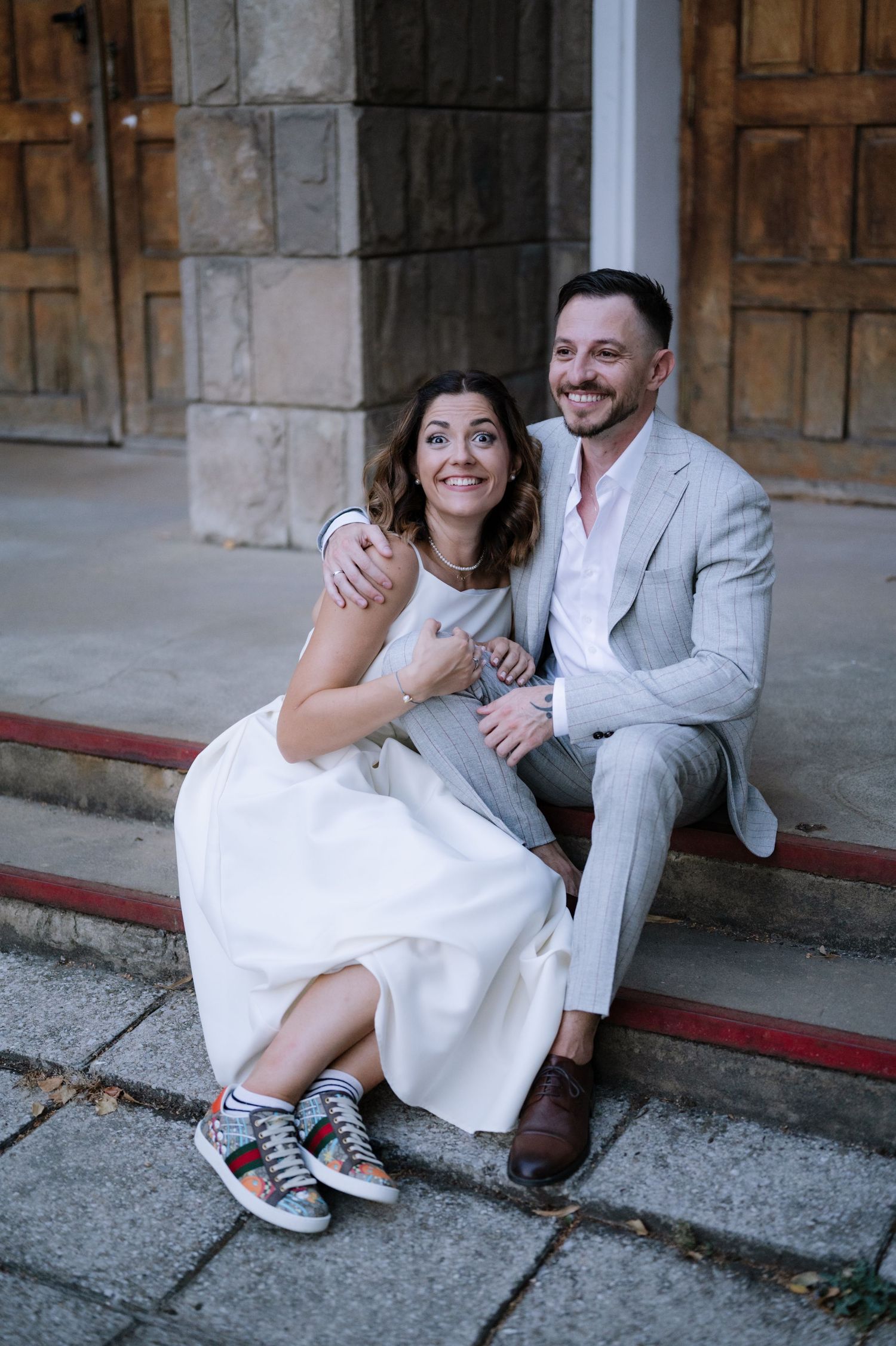 A couple sits on stone steps together, the bride in a white dress and sneakers, groom in light gray suit, sharing a joyful moment.