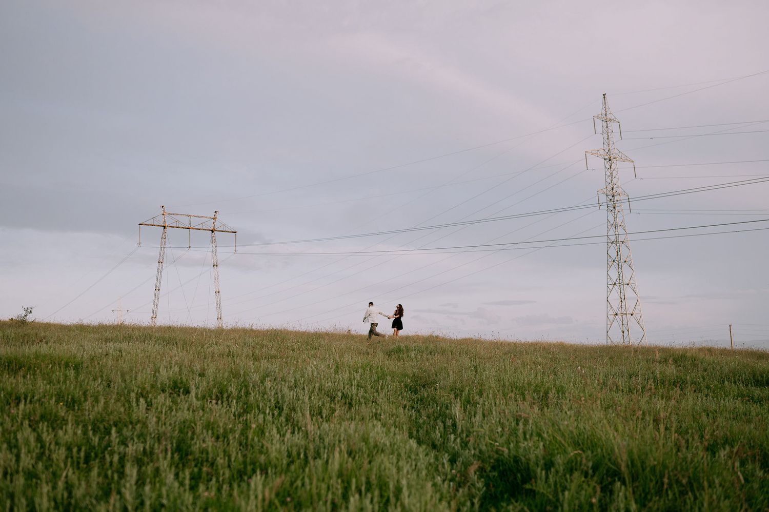 Distant figures traverse a green hillside dotted with electrical towers under an overcast sky.