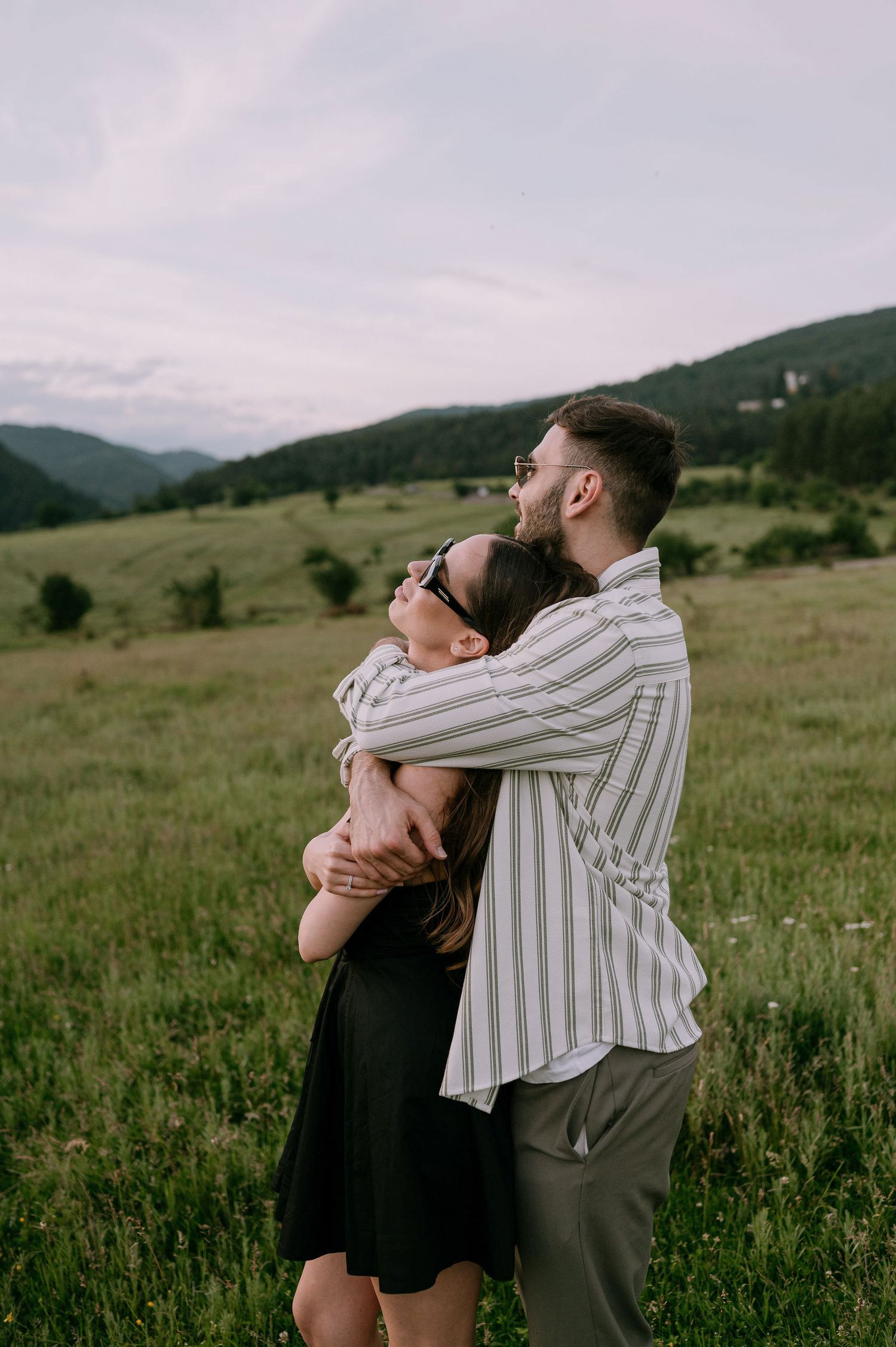 A couple embraces in a meadow with rolling mountains and green hills in the background during golden hour.