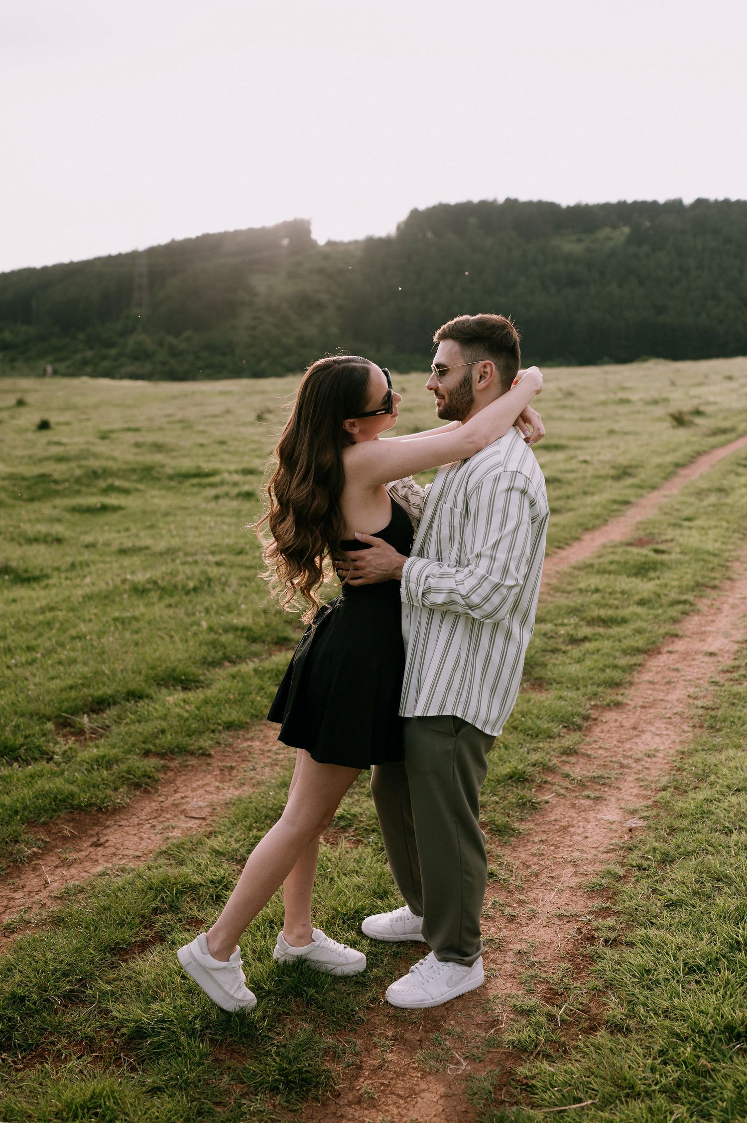 A couple shares a romantic moment on a dirt path through green rolling hills at sunset.