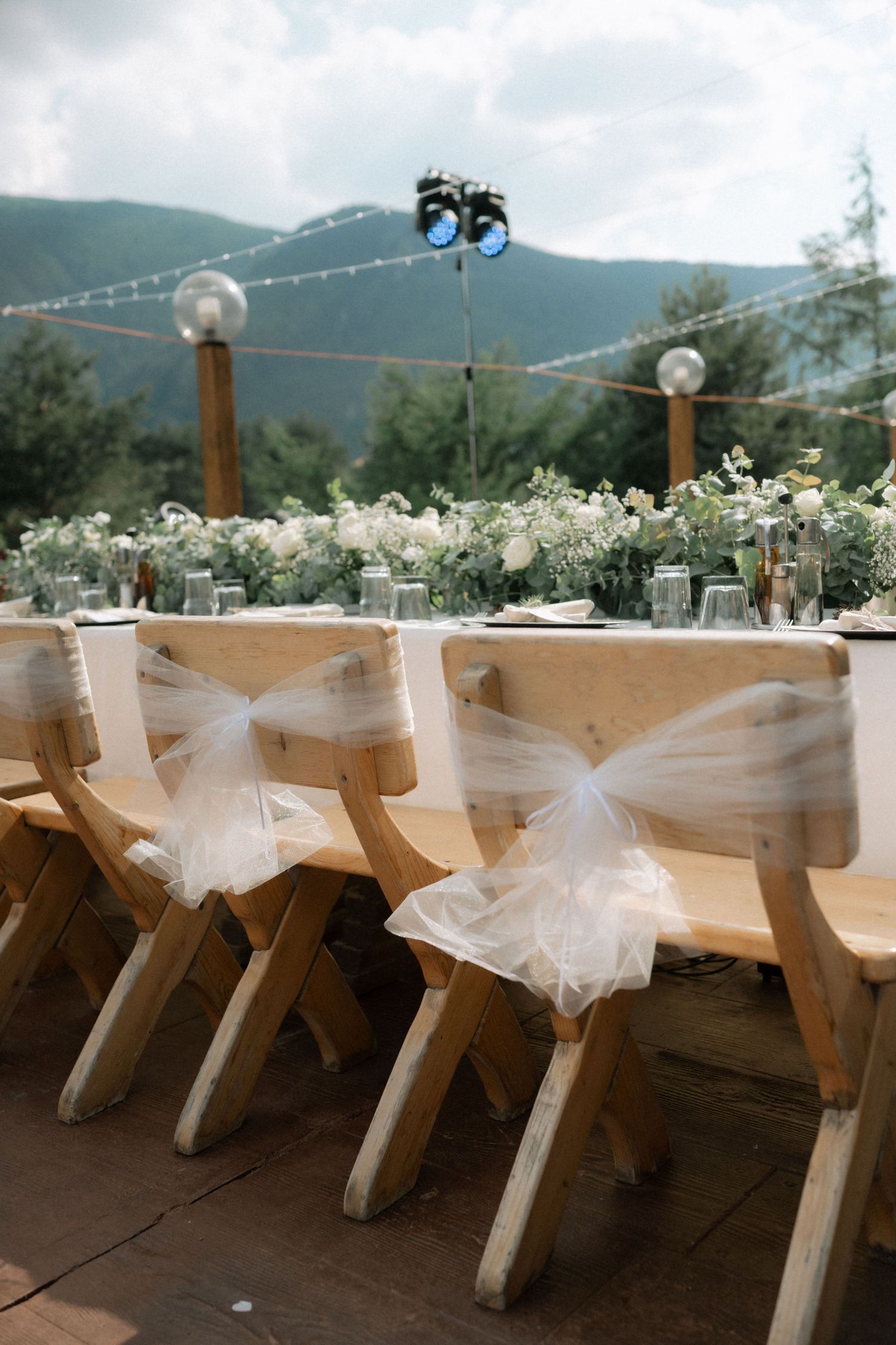 Wooden chairs decorated with white tulle bows at an outdoor mountain wedding reception with white floral centerpieces.