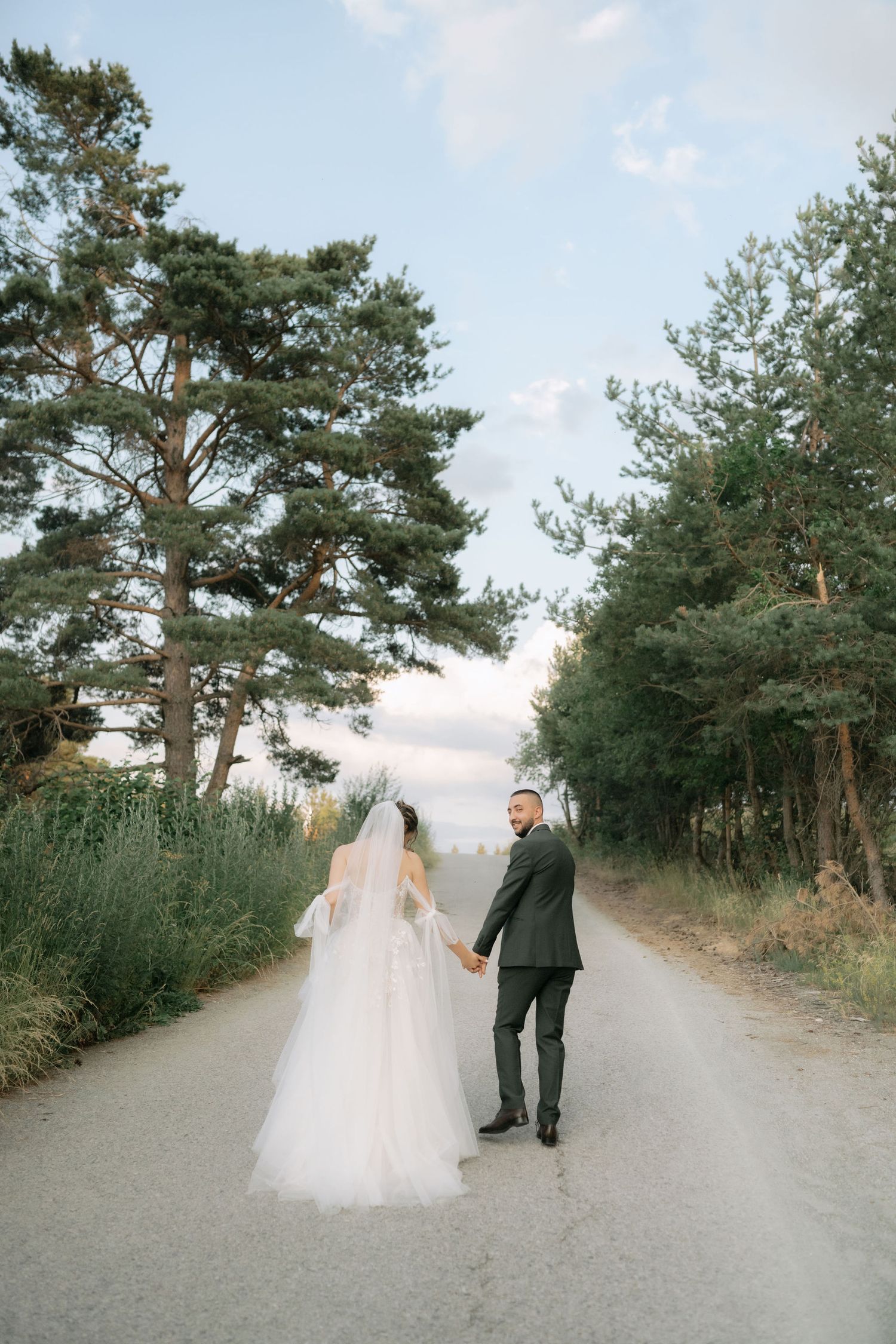 A couple walks down a serene forest path lined with tall pine trees on their wedding day.