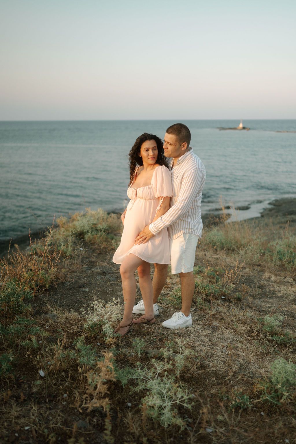 A couple shares a romantic moment on a coastal cliff overlooking the ocean at sunset.