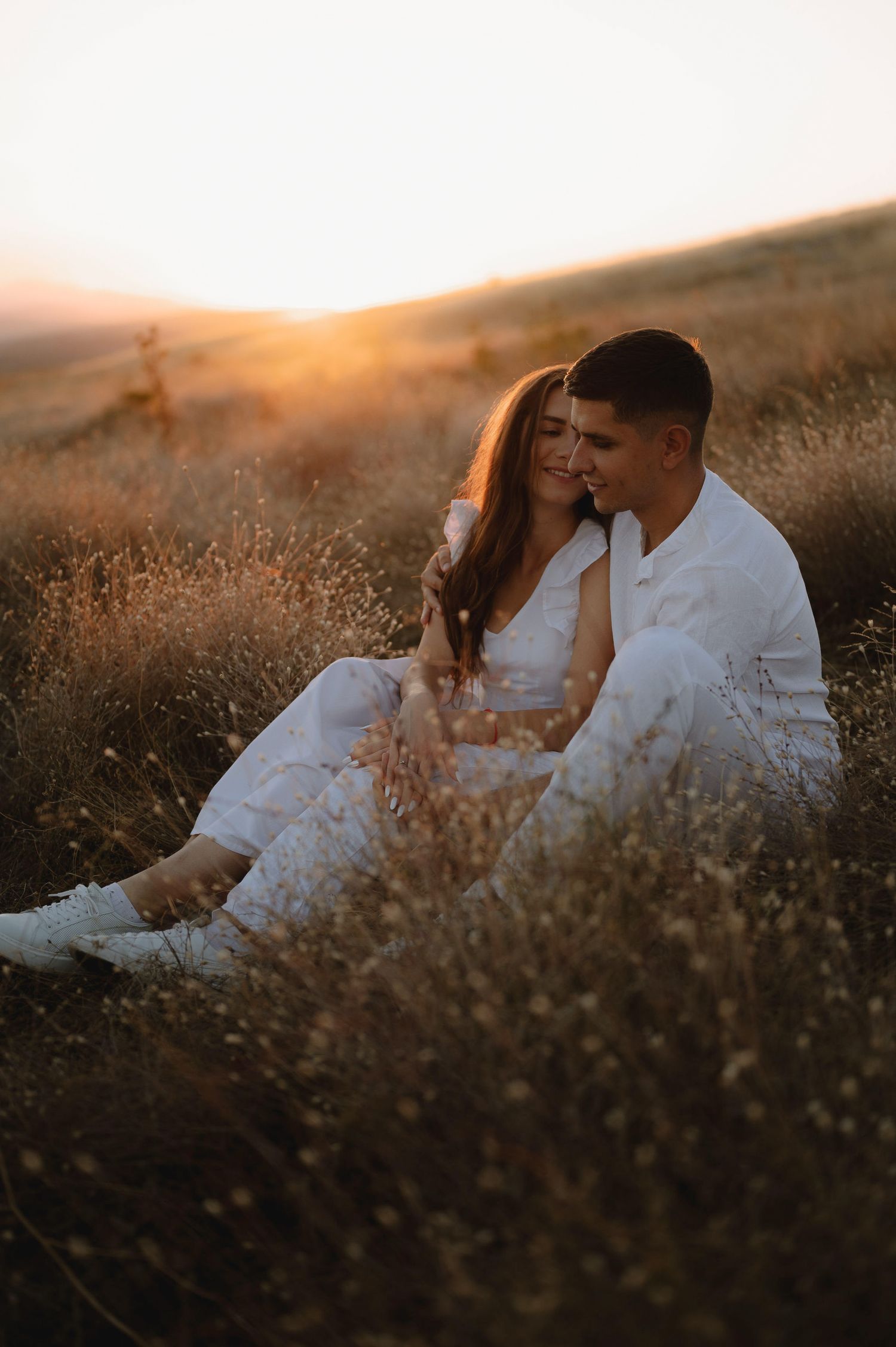 A couple sits together in a golden field during sunset, embracing amid tall dry grass and warm glowing light.