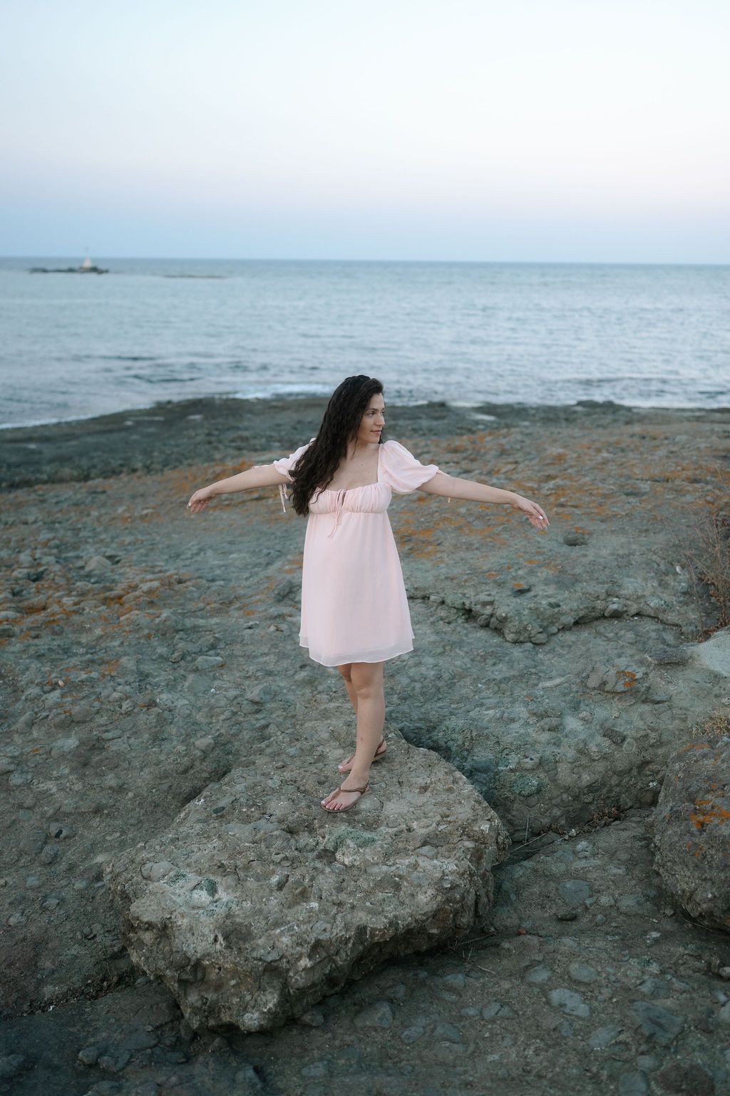 A person in a white dress balances gracefully on rocky coastal formations at sunset near the ocean.
