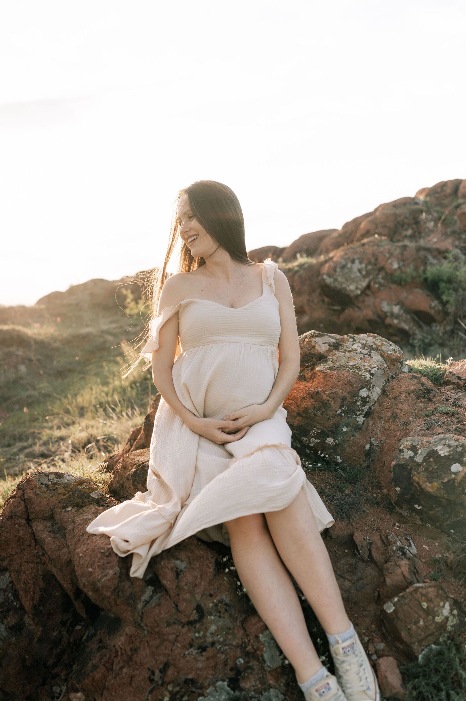 A person in a white dress sits on coastal rocks during golden hour as sunlight creates a dreamy atmosphere.