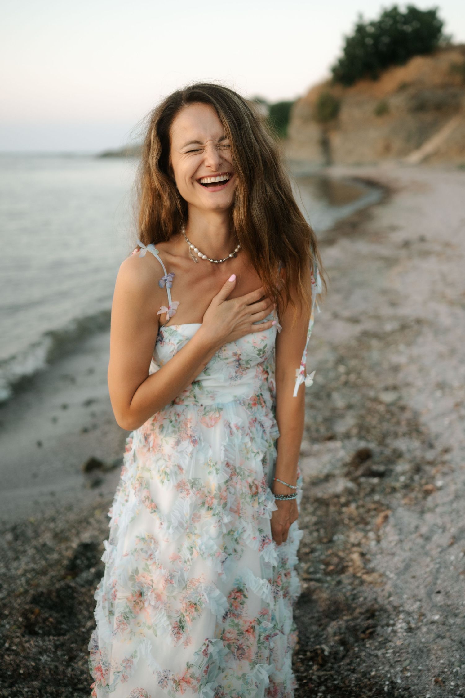 A person in a floral dress laughs joyfully while walking along a rocky beach at sunset.