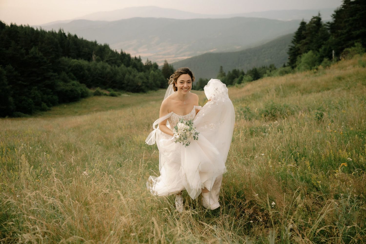 A flowing white wedding dress billows in the mountain breeze as wildflowers sway on a scenic hillside meadow.