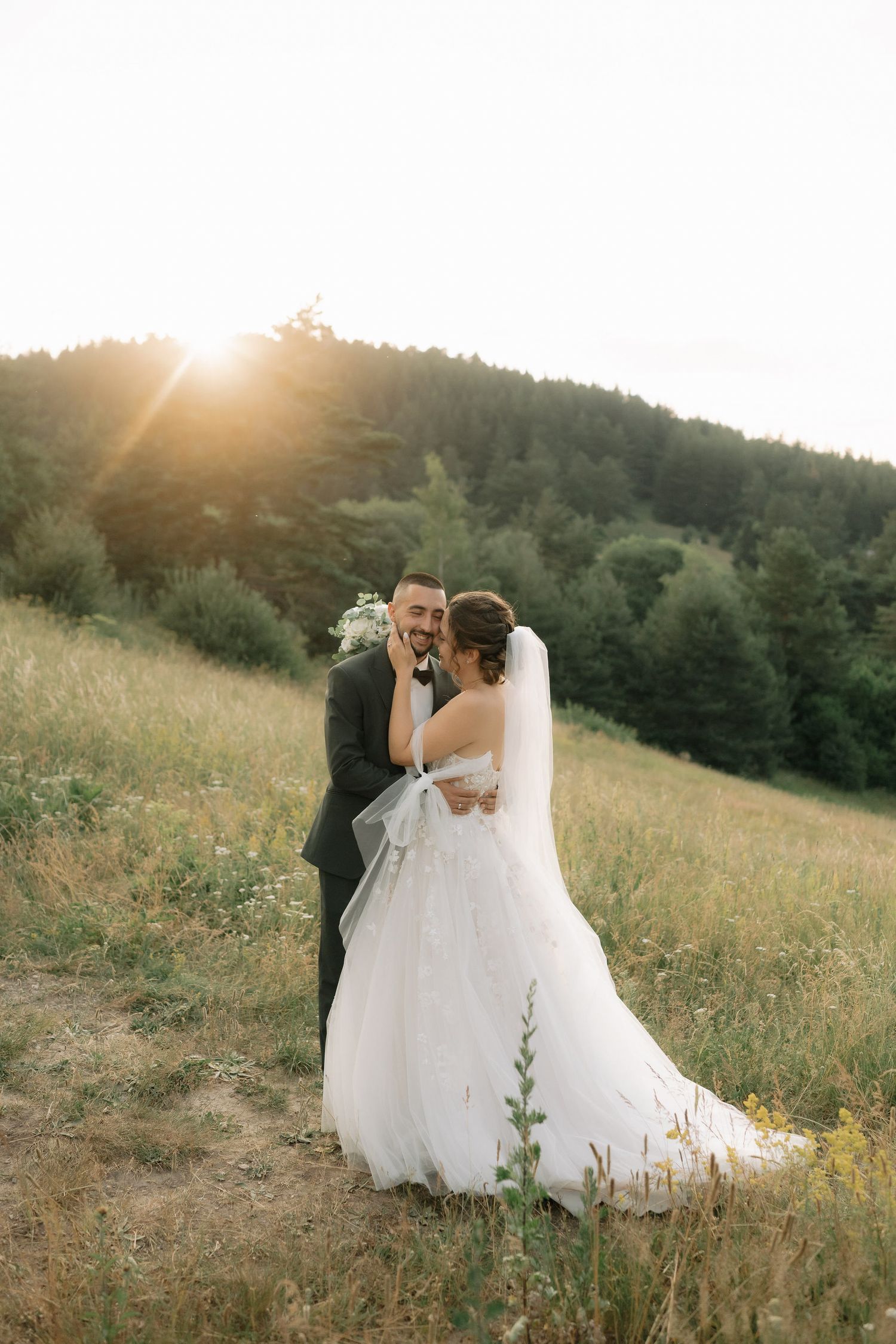 A romantic wedding couple embraces in a sun-drenched meadow with mountains and forest in the background at golden hour.
