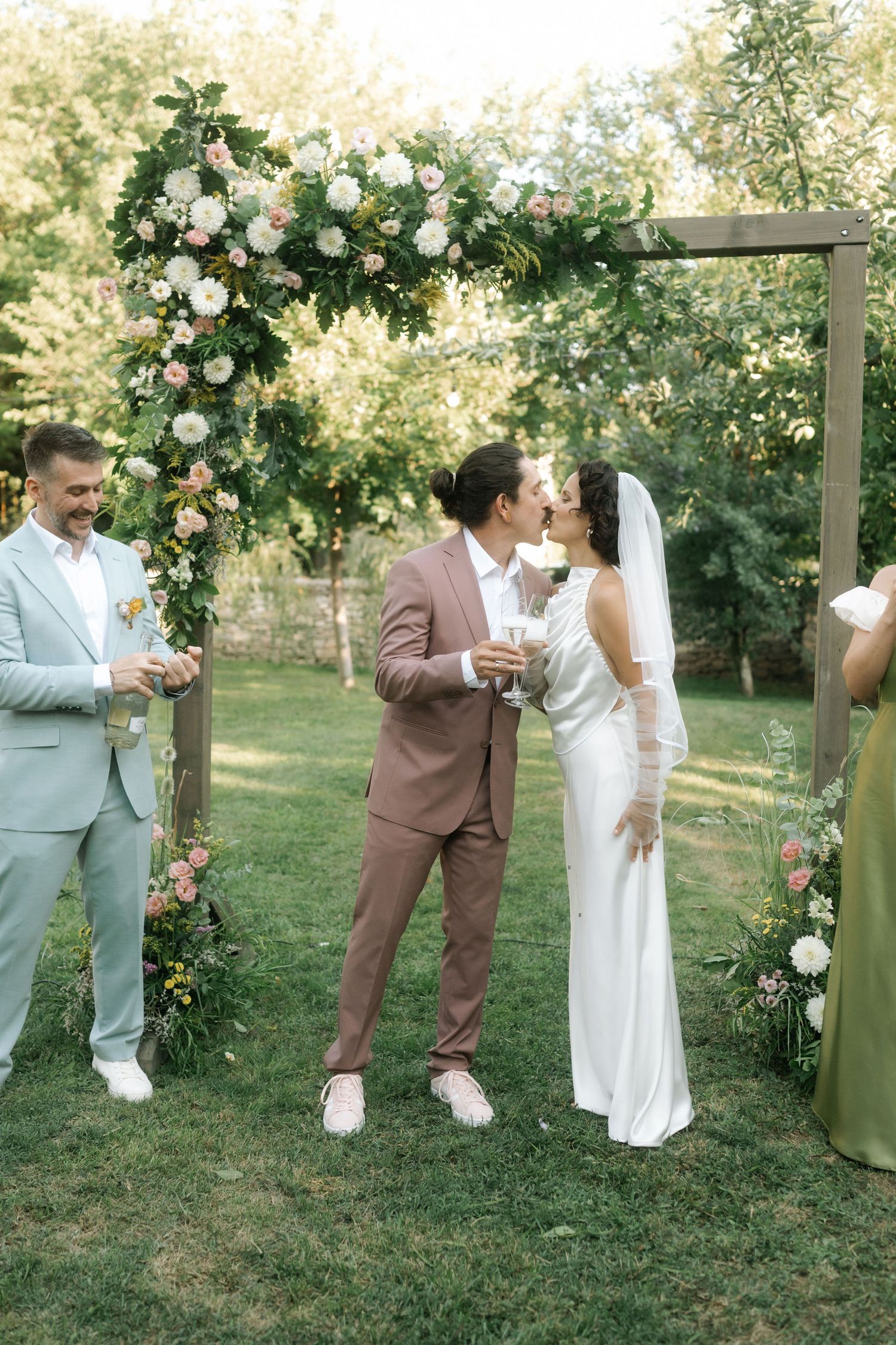 Wedding party poses together in a garden setting with white floral decorations and natural greenery.