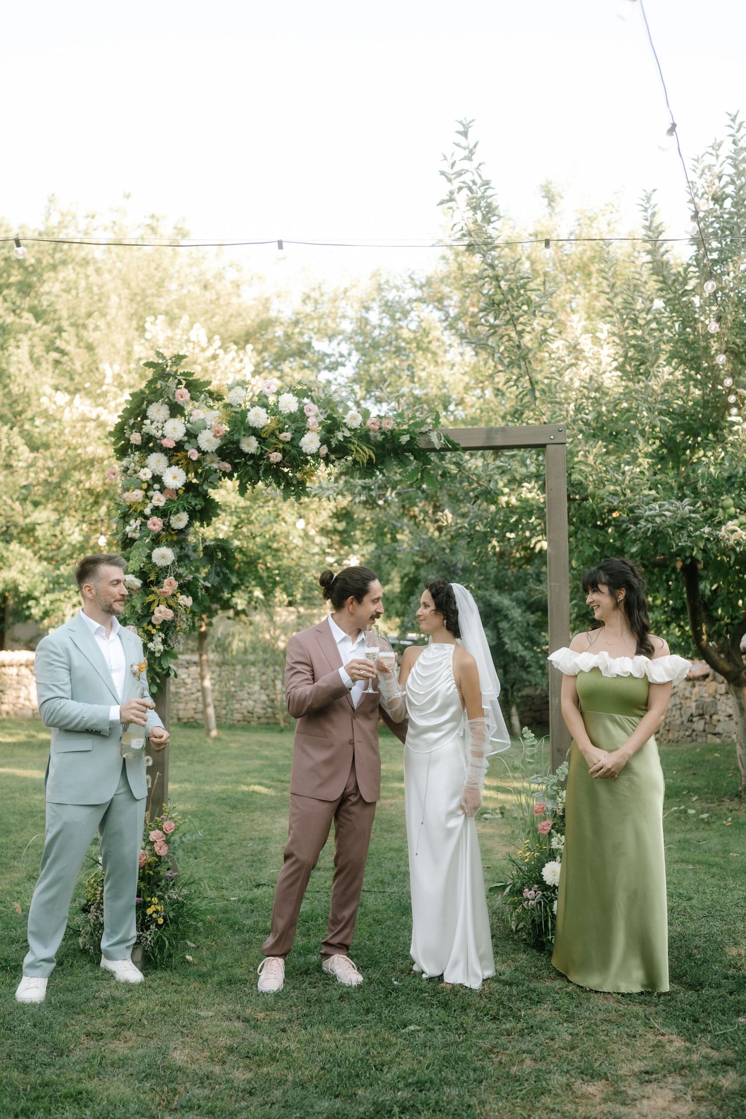 A wedding party in green and brown attire poses together in a garden setting with greenery and trees in the background.
