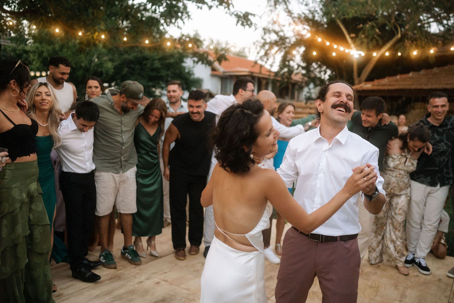 A couple shares their first dance at an outdoor wedding reception under string lights while guests watch and celebrate.