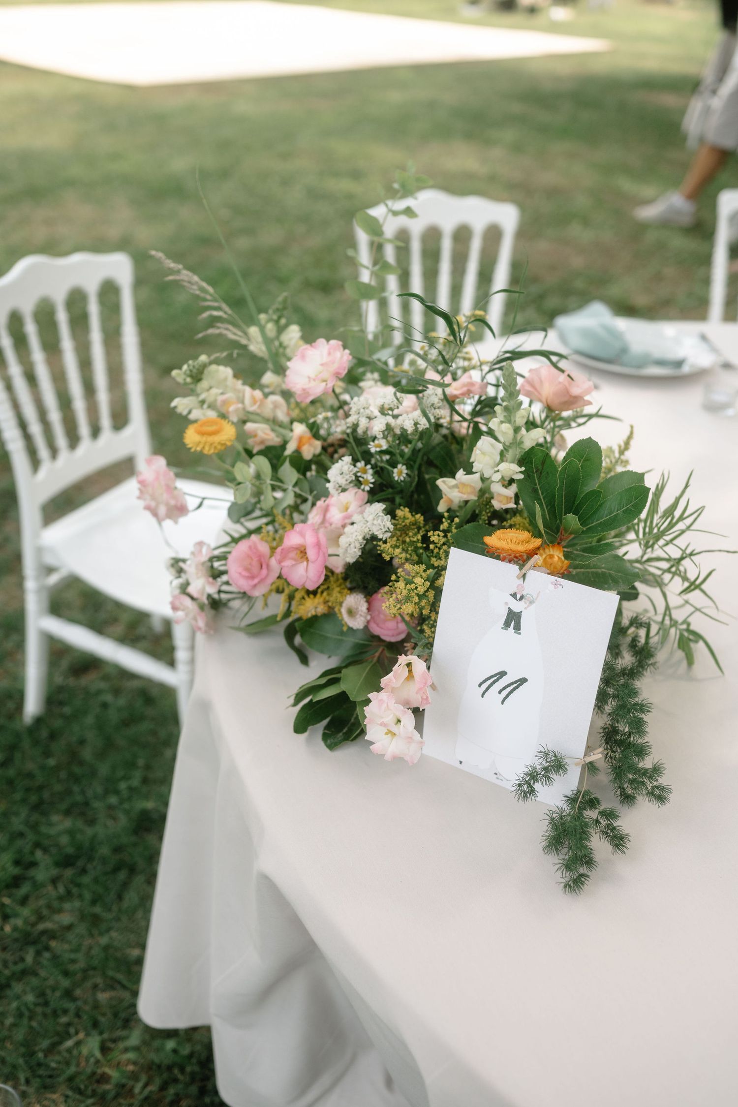 White chairs at an outdoor wedding reception decorated with pink and orange floral arrangements on white tablecloths.