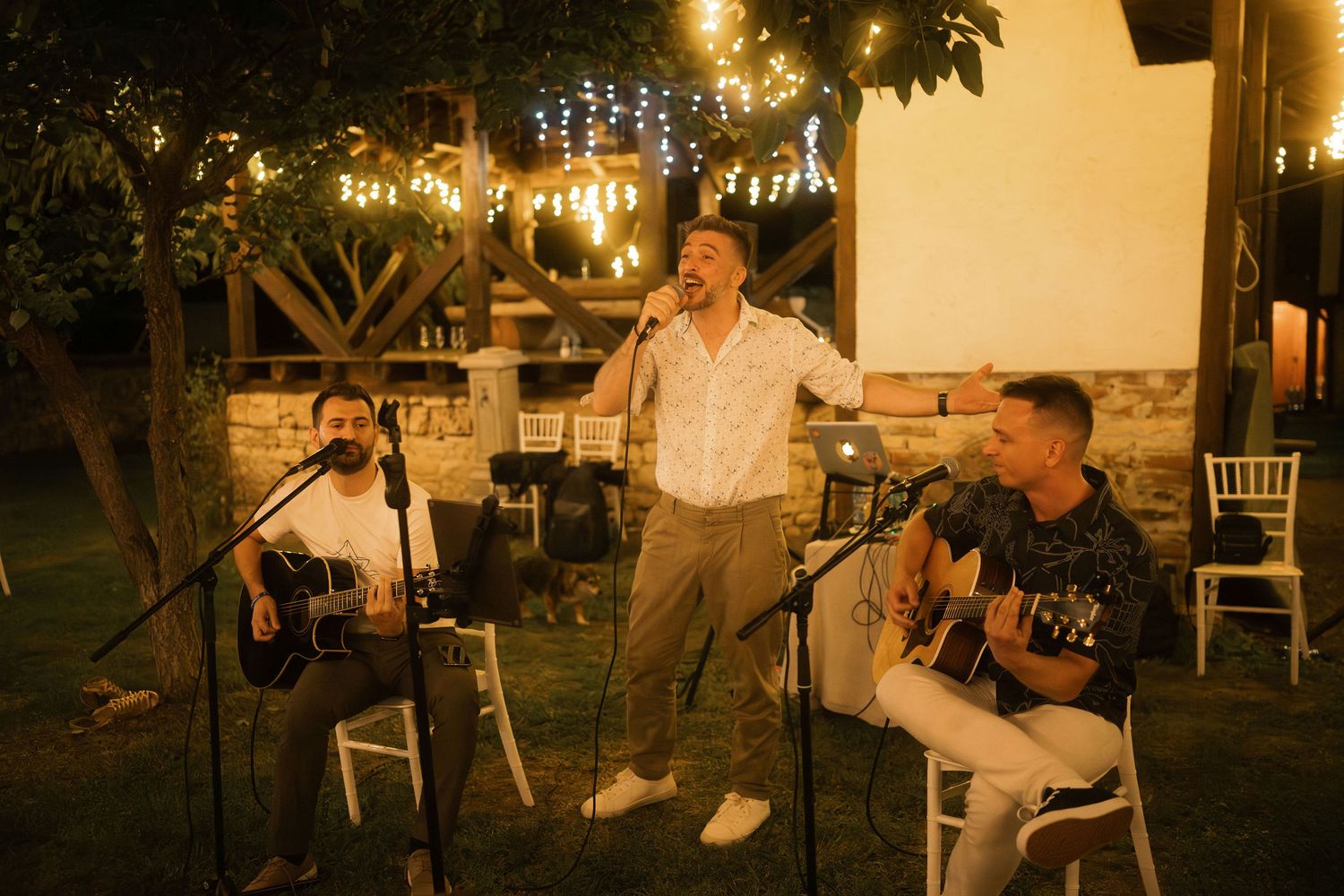 String lights illuminate an outdoor evening wedding reception with guests seated at tables in a garden setting.
