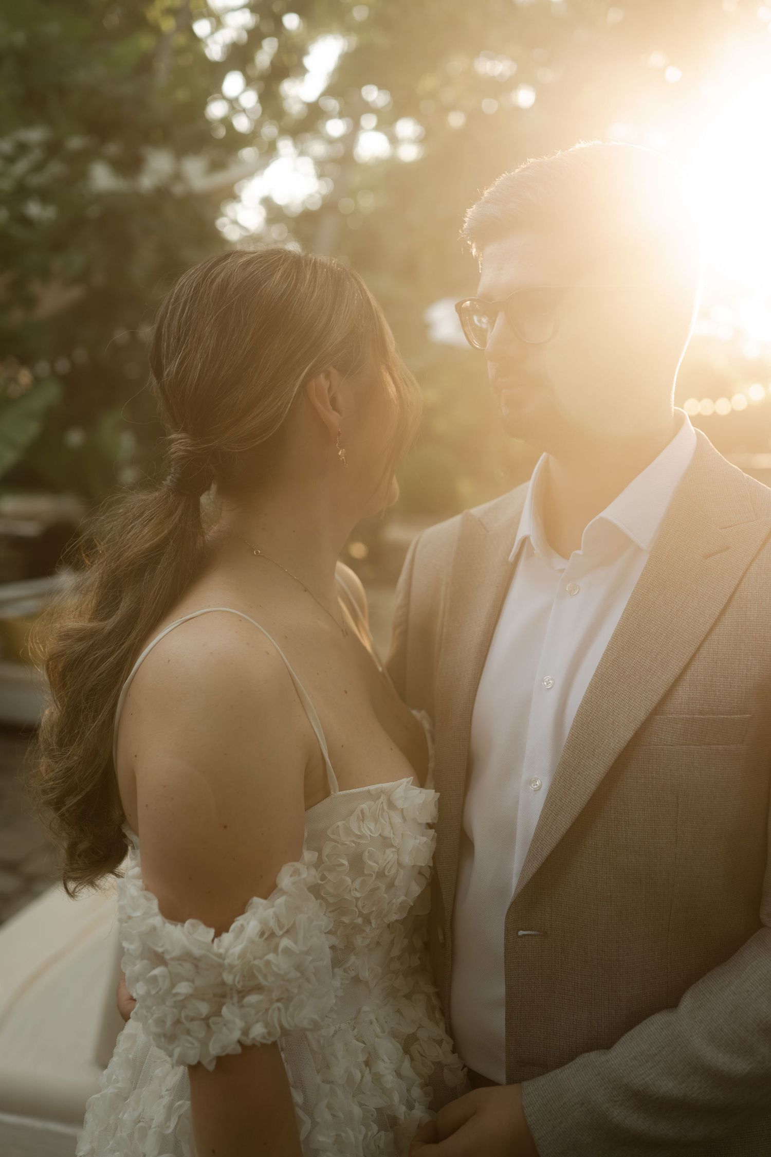 A romantic wedding couple shares an intimate moment in golden sunlight with soft bokeh through trees.