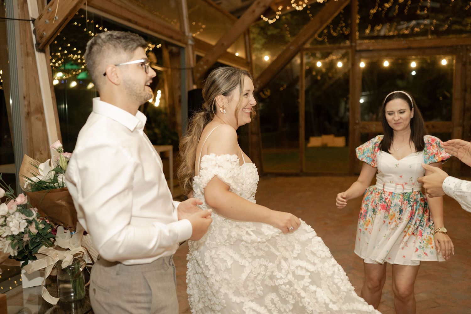 Wedding guests dance and celebrate together on a rustic wooden dance floor illuminated by string lights overhead.