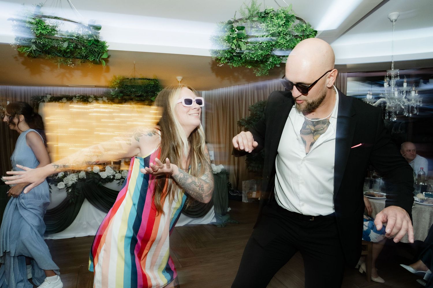 Two people dance energetically on a wooden floor at an indoor event with decorative plants visible in the background.