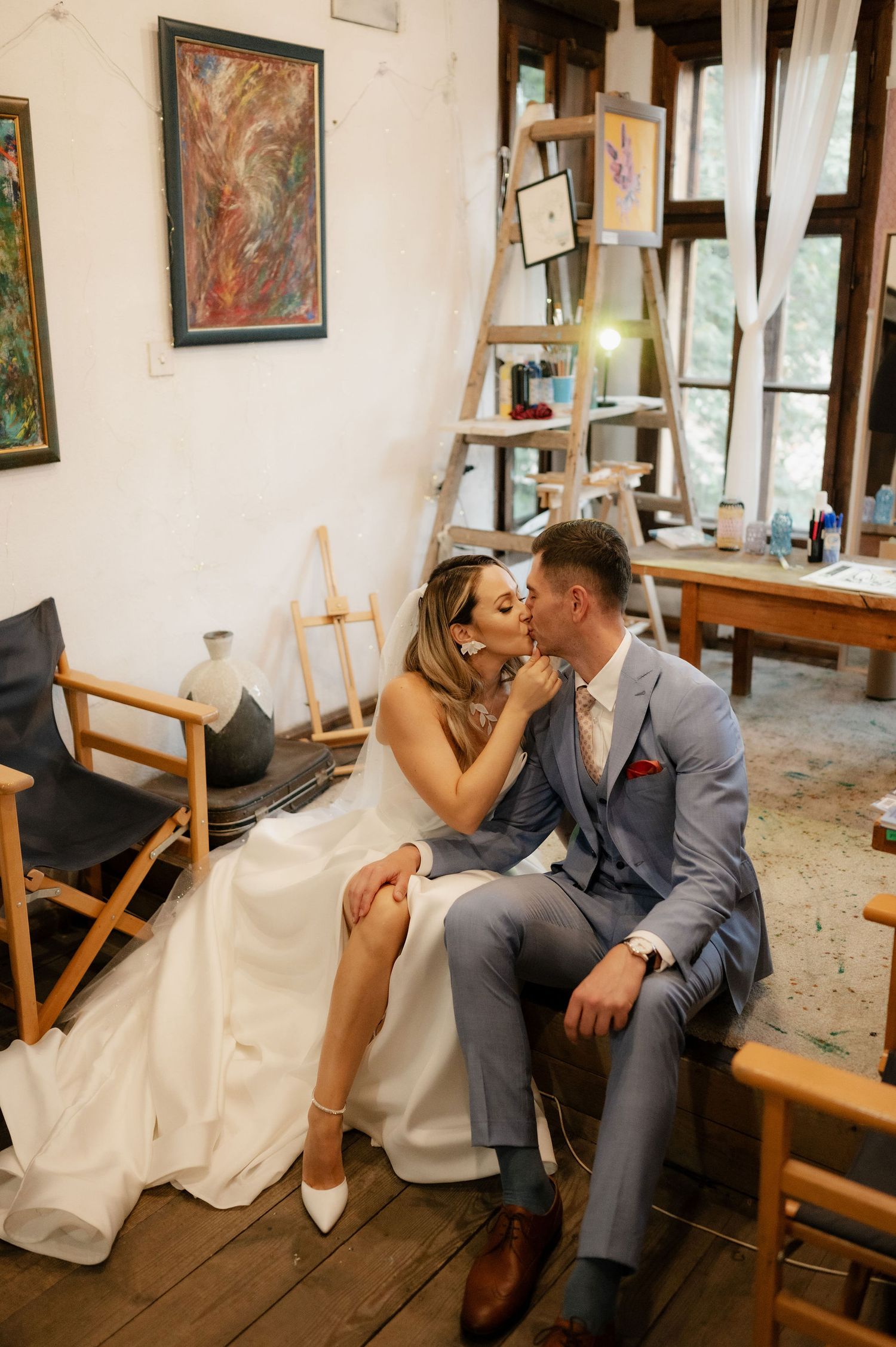 Newlyweds share tender moments while sitting on wooden chairs in an art-filled room.