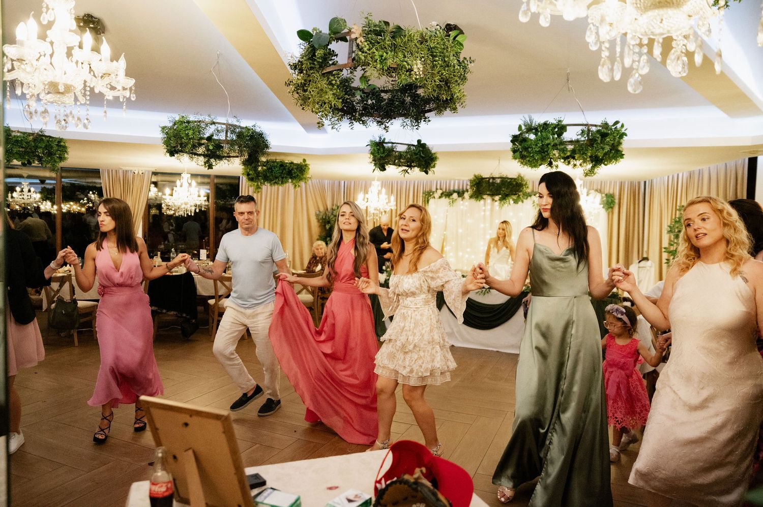 Wedding guests dance and celebrate on an illuminated dance floor under decorative lights at an indoor reception venue.