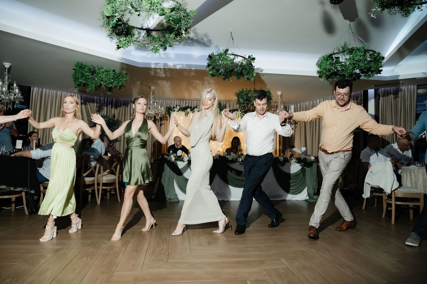 Dancers in light colored outfits perform an energetic line dance routine at an indoor reception venue.