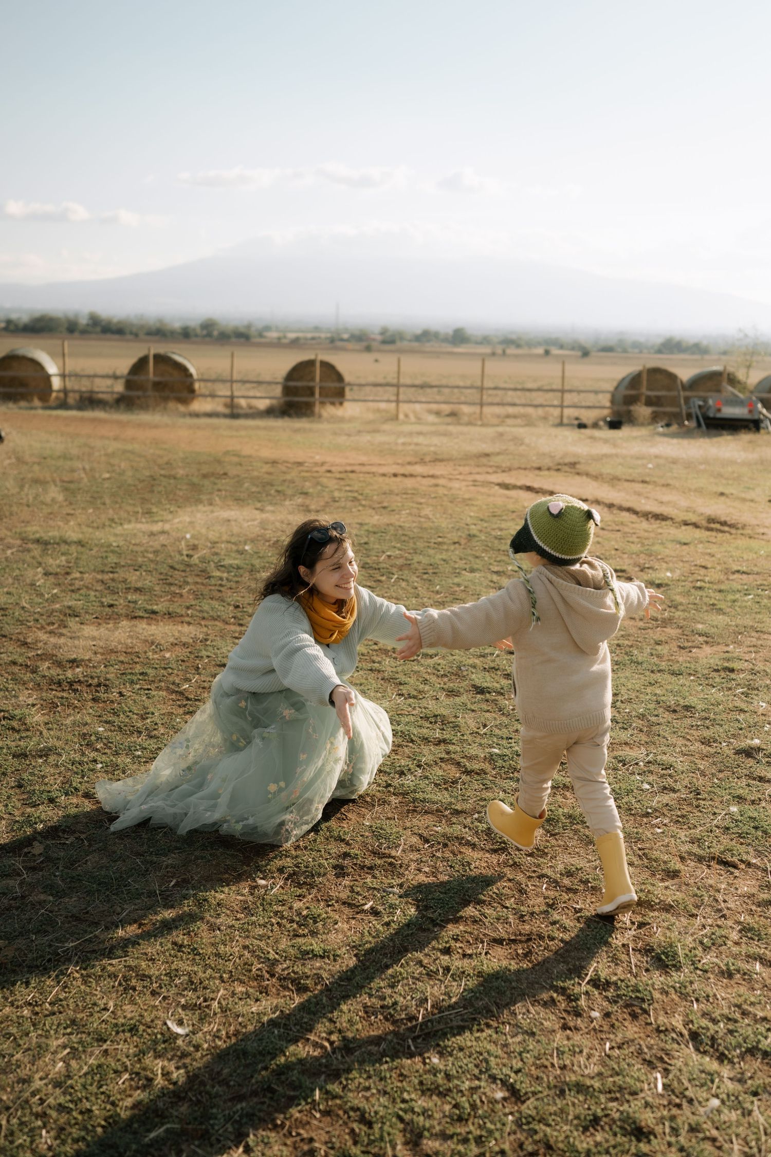 A couple in wedding attire playfully runs through an open field at sunset casting long shadows on the grass.