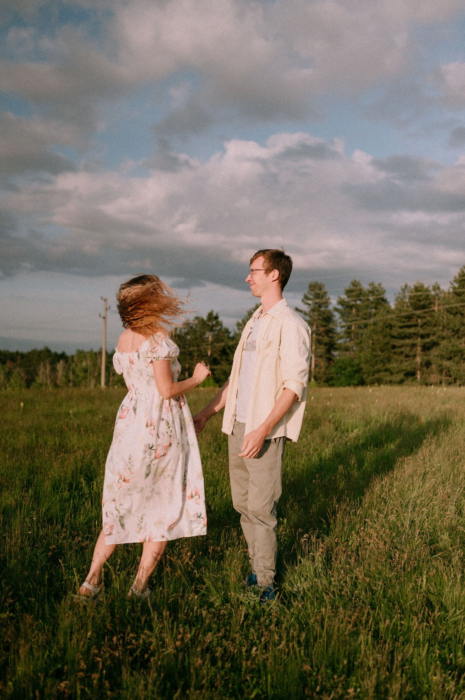 Couple dancing in a meadow with pine trees and cloudy sky in the background.
