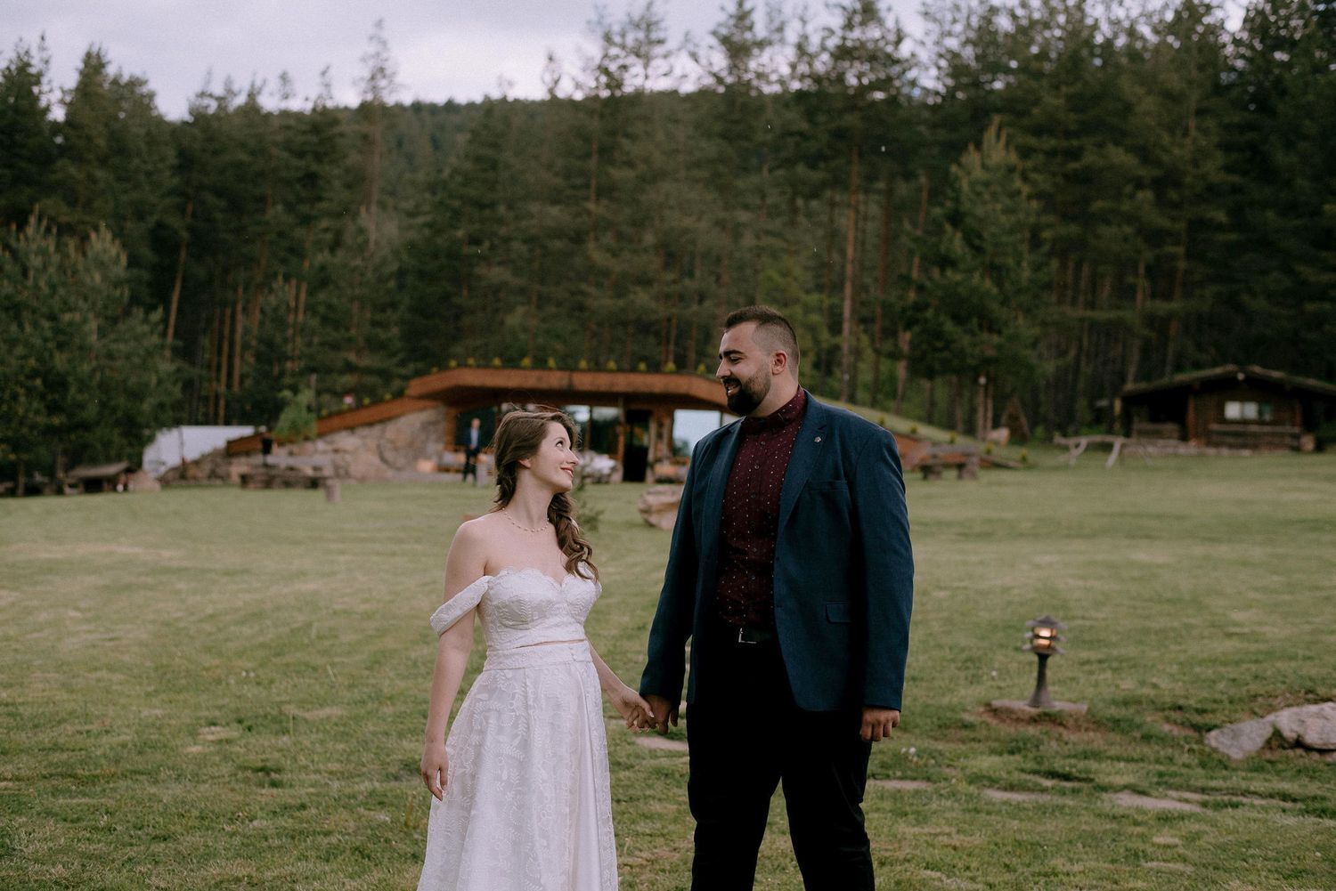 A couple walks hand in hand on a grassy slope with wooden cabins in the background.
