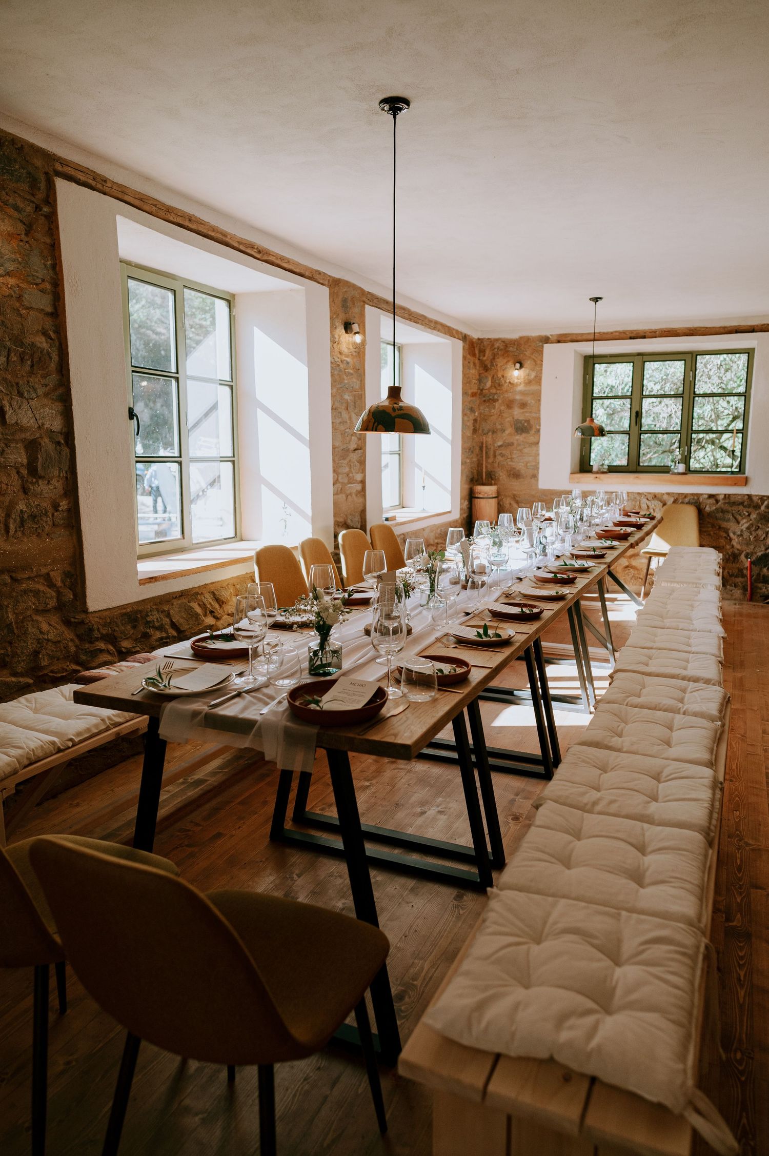 Long dining table with tufted bench seating and modern chairs in stone-walled room with natural light.
