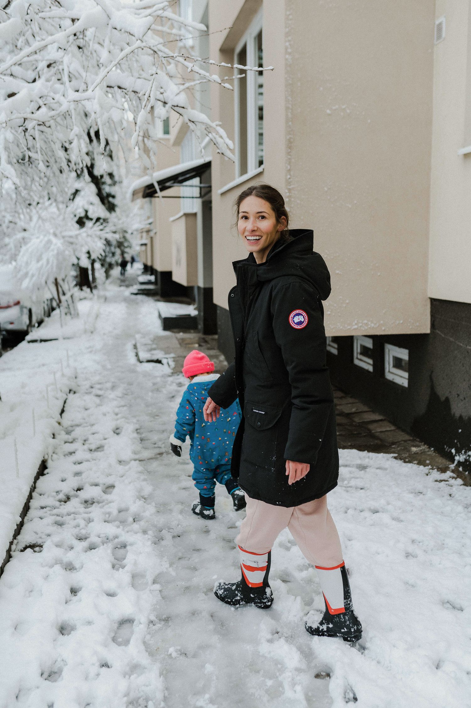 A winter scene with a person in a black Canada Goose coat and a small child walking through snow-covered ground.