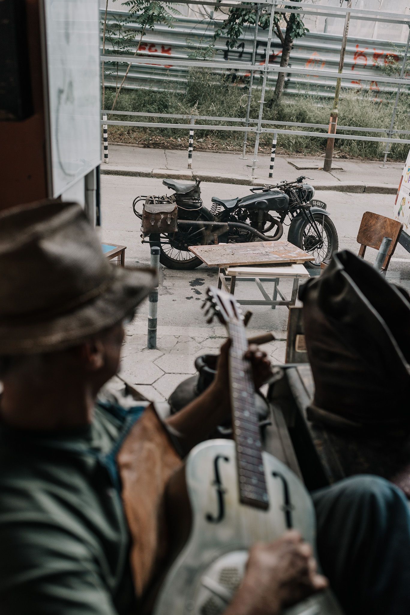 Person in green jacket and hat sitting by window with guitar.