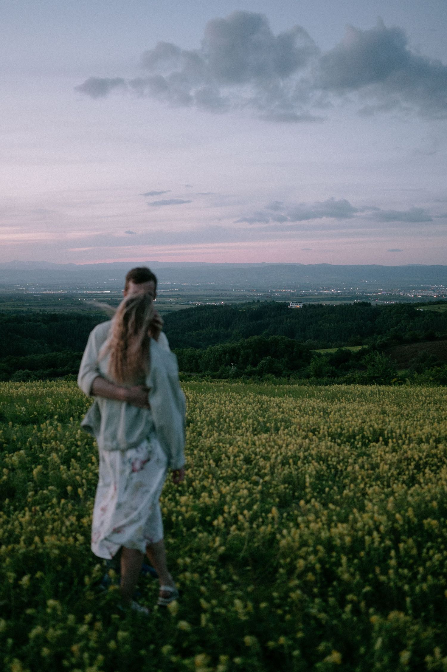 Two figures embrace while standing in a vast field of yellow wildflowers under moody skies.