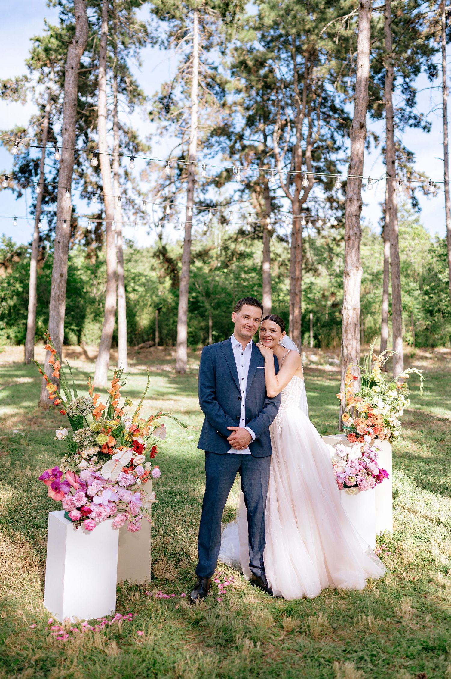 A romantic wedding photo featuring a couple surrounded by tall trees and colorful floral decorations.