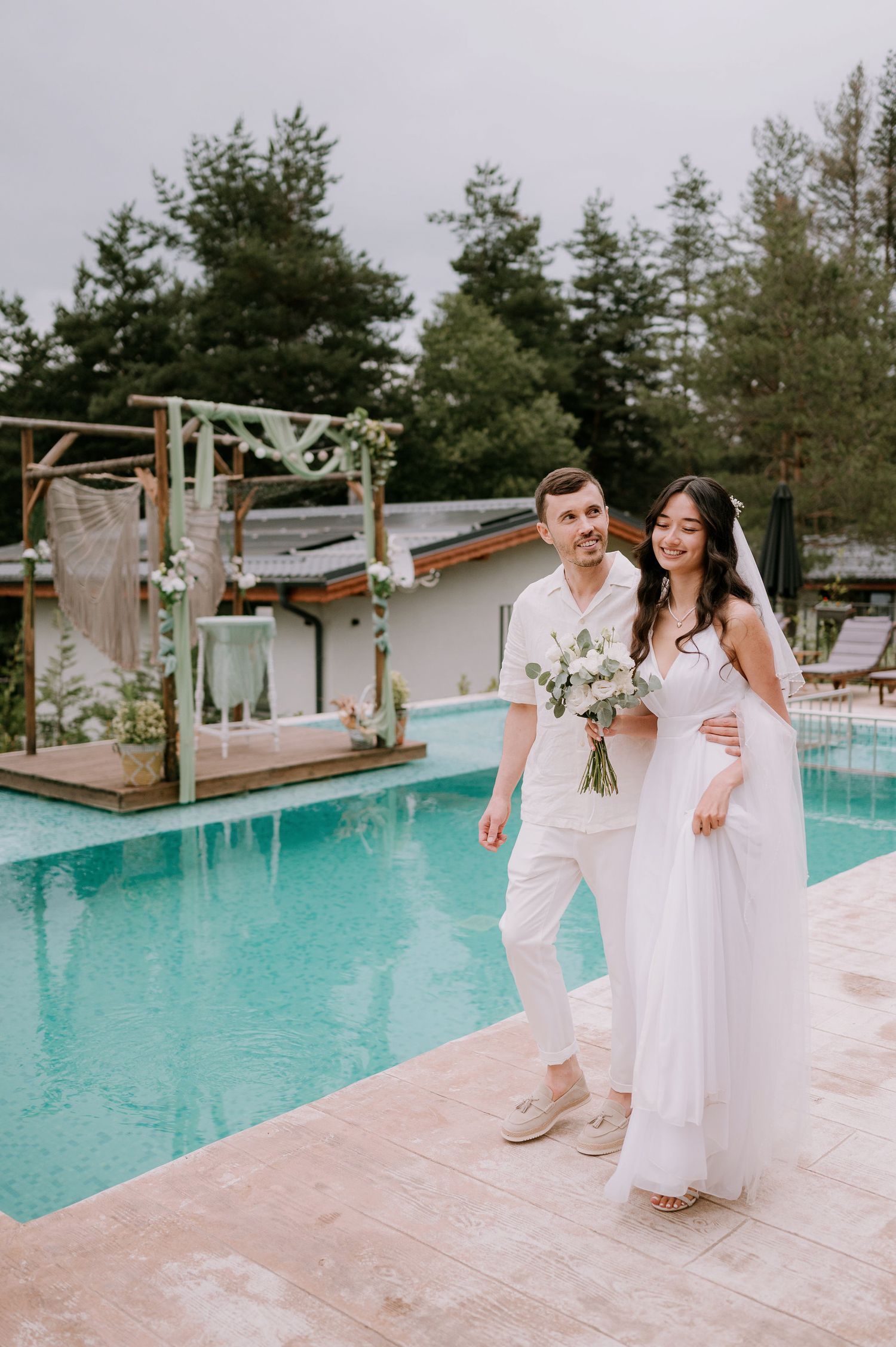 A couple in white attire shares a romantic moment by a turquoise pool surrounded by evergreen trees.