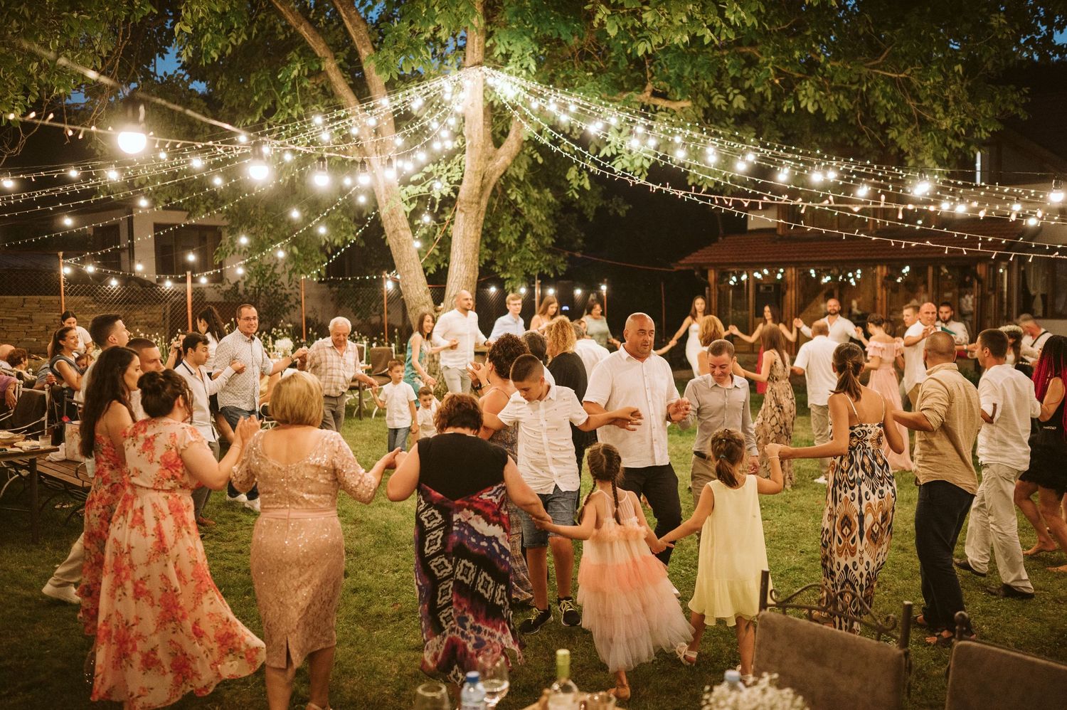 Outdoor wedding reception with guests in floral dresses dancing beneath string lights.