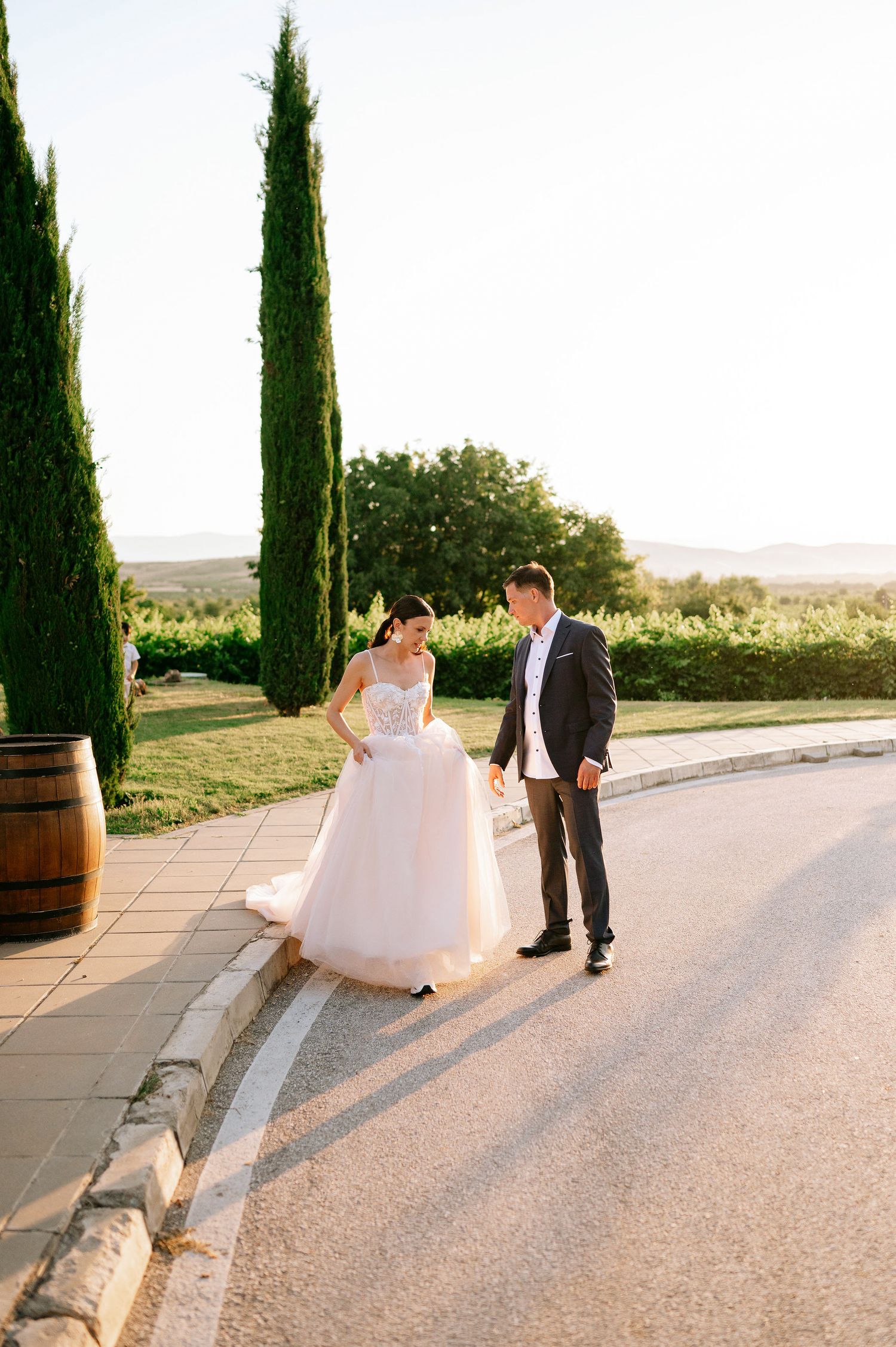 Newlyweds walking together on a sunlit path lined with cypress trees.