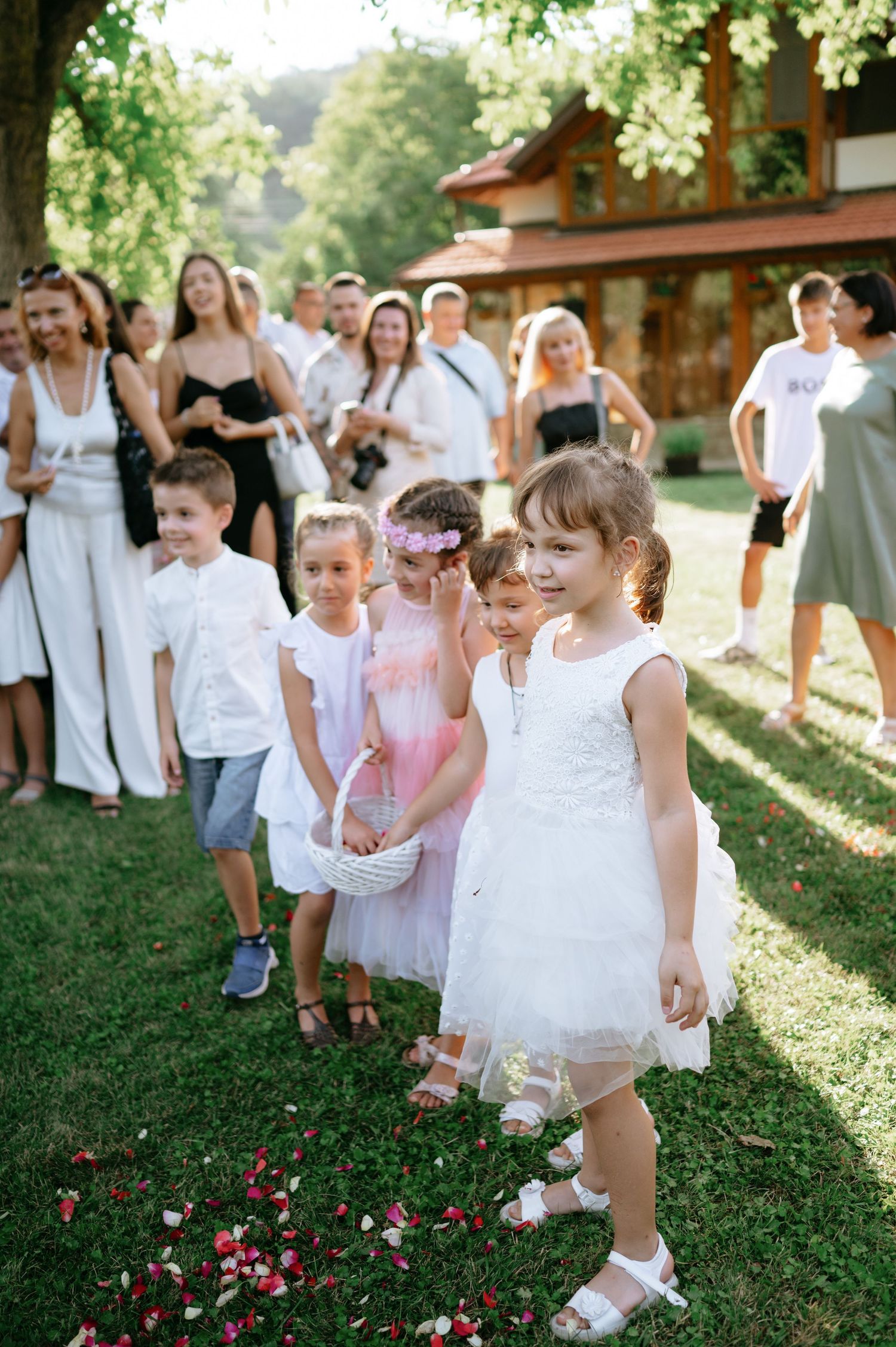 Young flower girls and children in white dresses walking on rose petal-covered grass at an outdoor wedding.