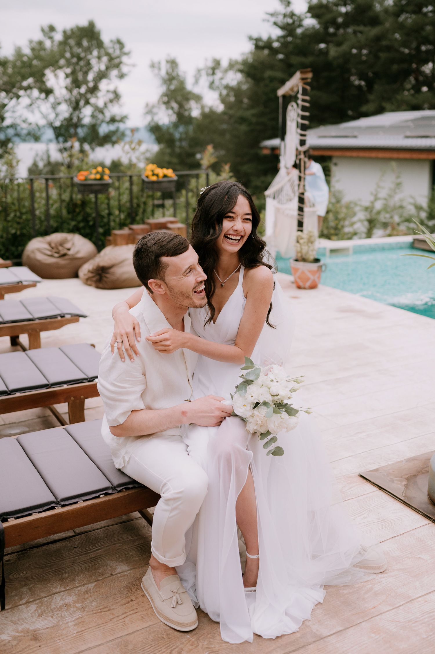 A couple shares a candid moment of laughter while sitting poolside in matching white outfits.