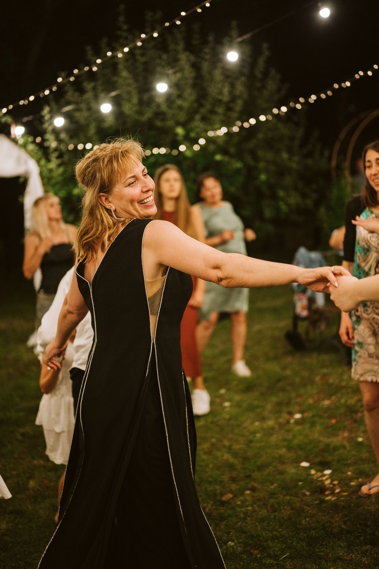A person in a black dress dances joyfully under twinkling outdoor lights.