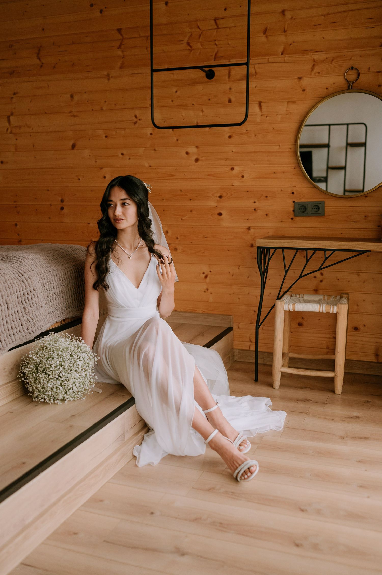 Ethereal bridal portrait in white dress sitting on wooden floor with baby's breath bouquet in rustic cabin setting.