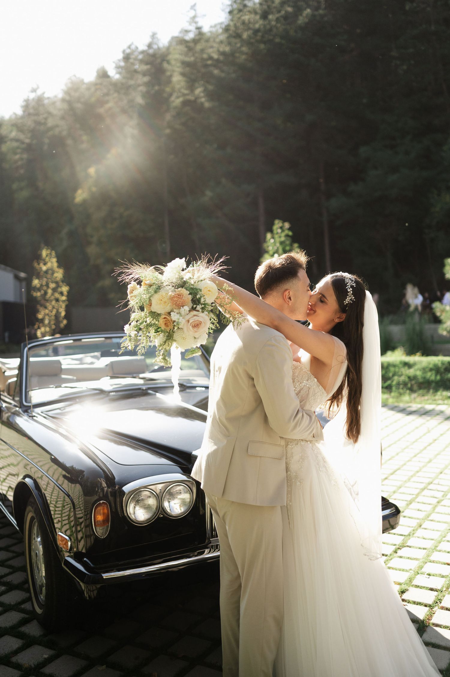 A joyful wedding celebration photographed beside a vintage automobile in natural sunlight.