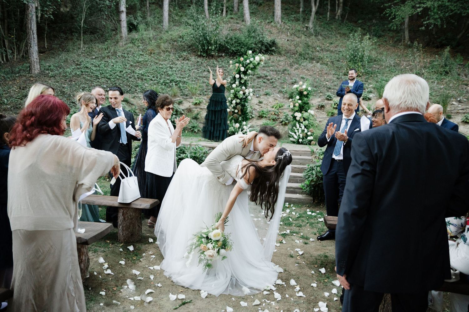 Guests celebrate at an outdoor forest wedding ceremony with white floral decorations.