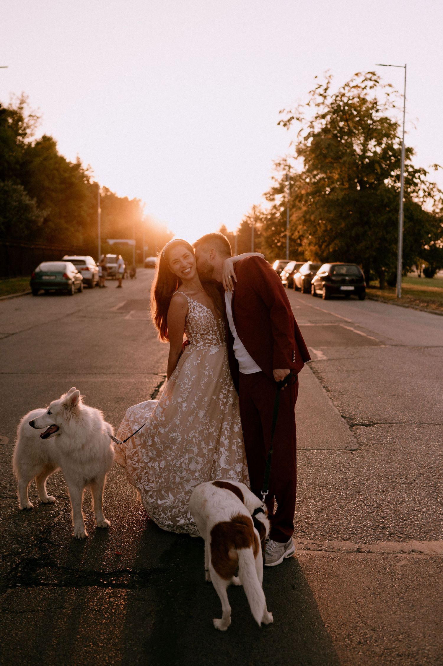 A couple in formal wear stands with their white dog on a parking lot during golden hour.