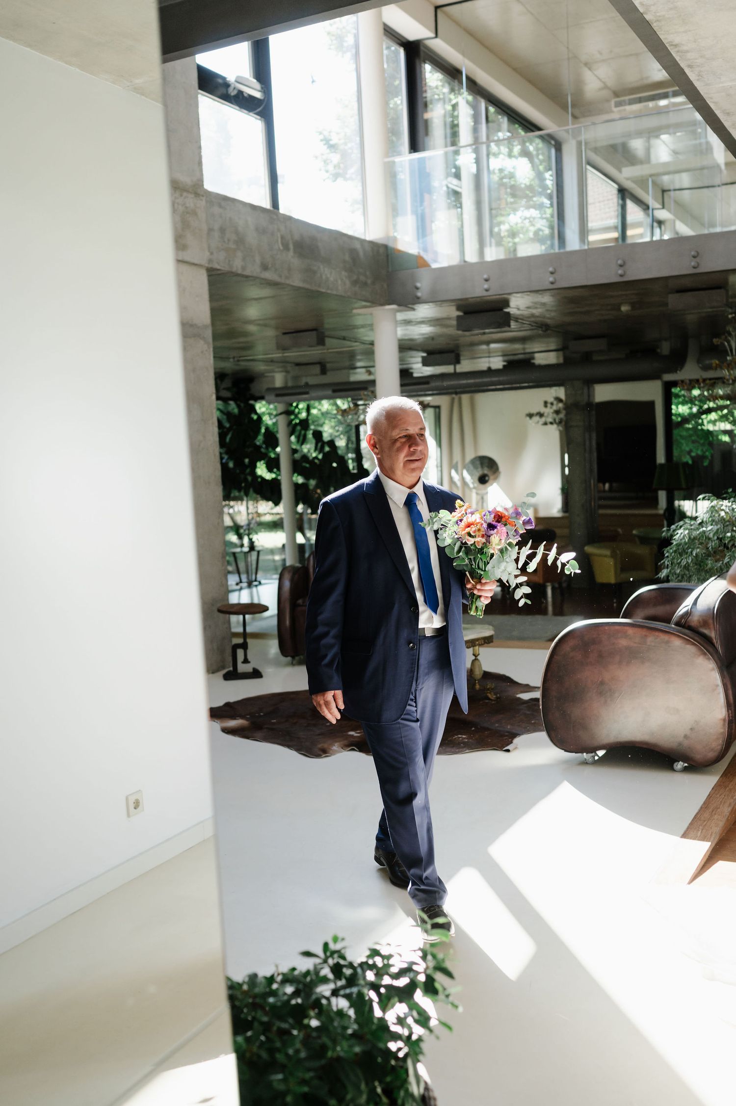 A person in a navy suit walks through a modern building interior with natural light streaming in.
