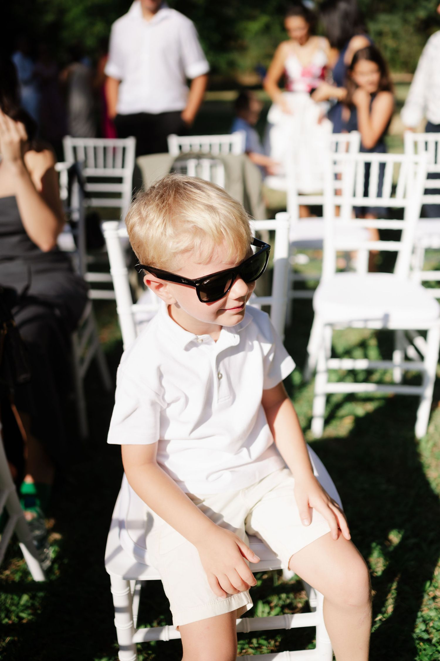 Young children in white outfits sit on white chairs at an outdoor event.