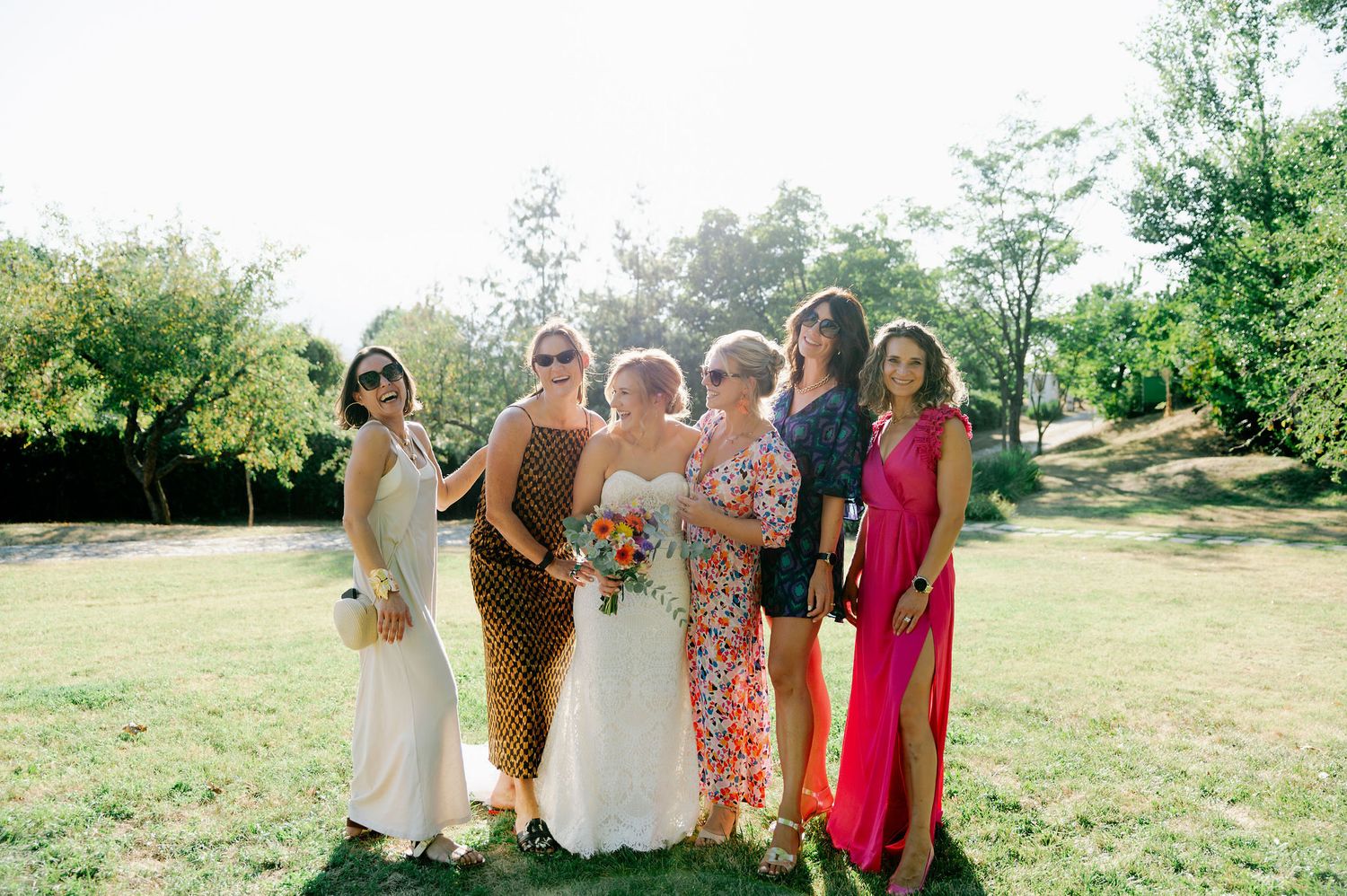 A group of wedding guests in colorful dresses pose together on a sunny grassy field.