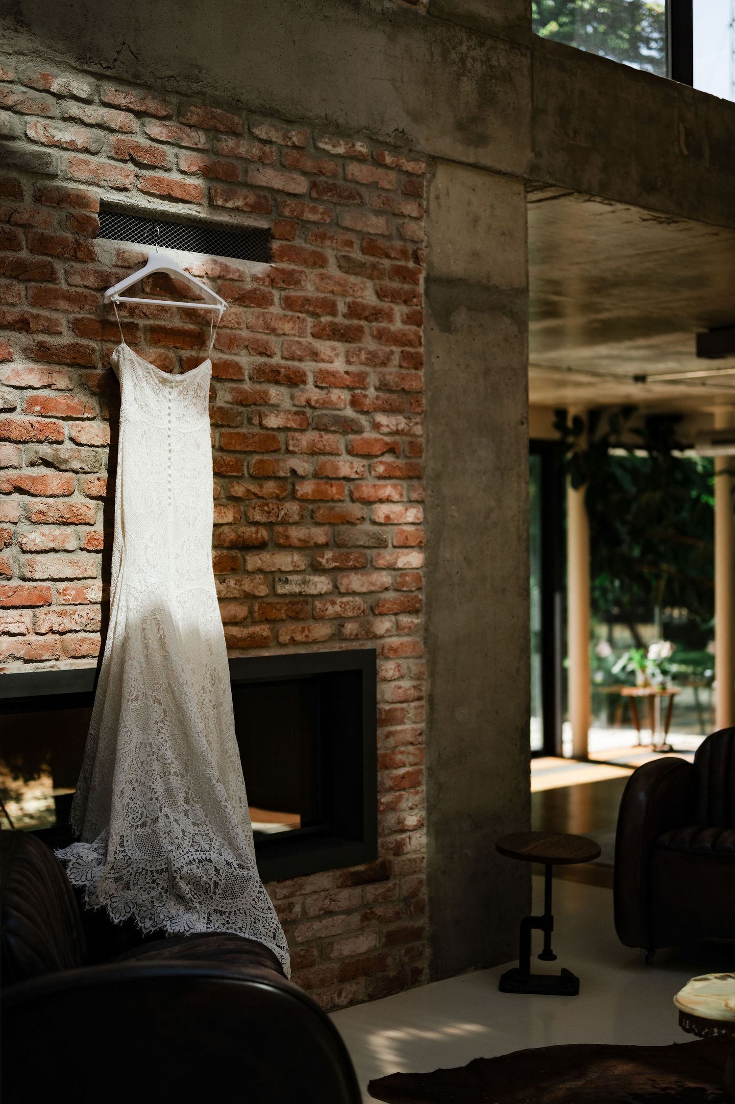 White lace wedding dress hanging against exposed brick wall in natural window light.