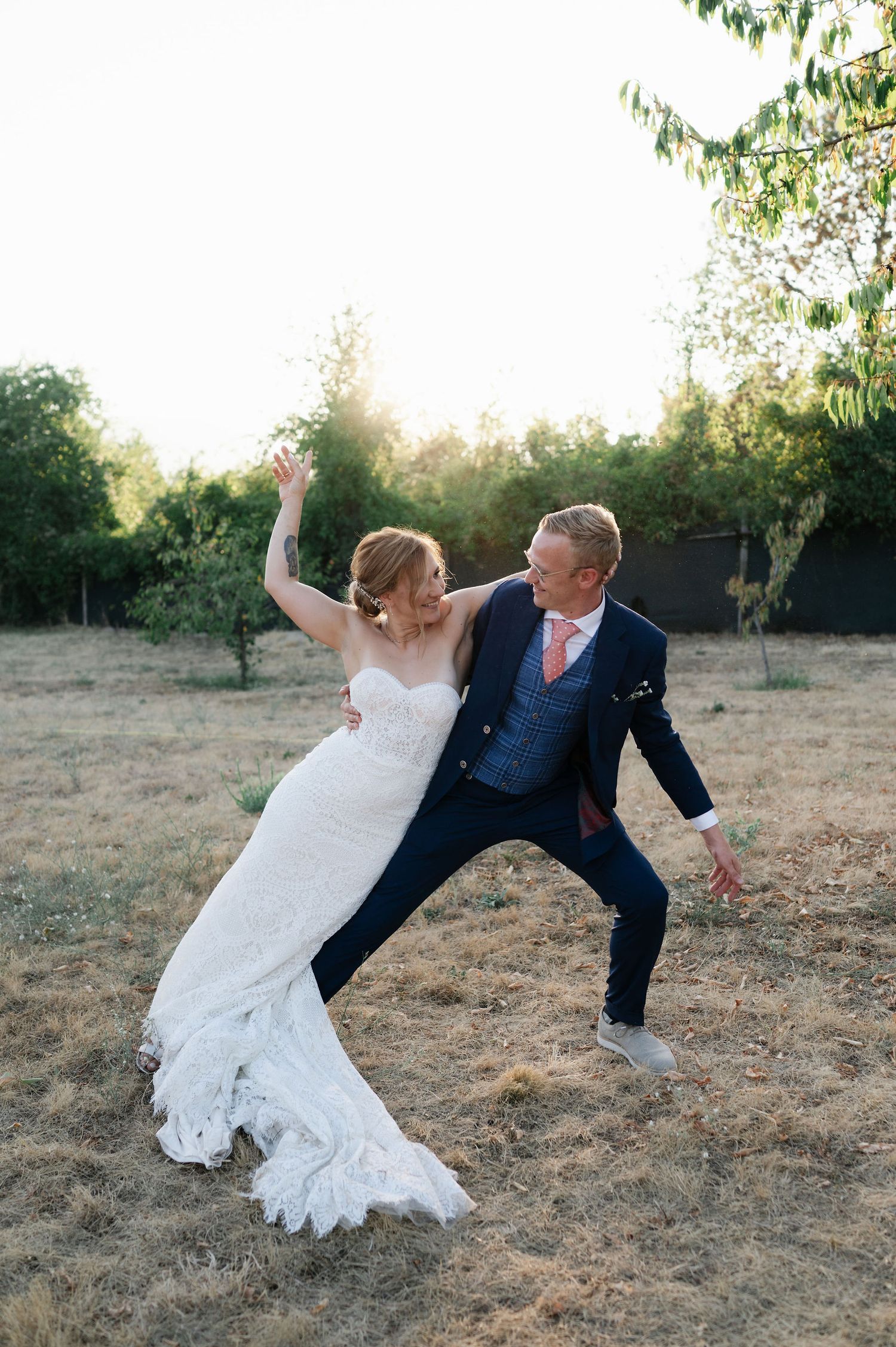 A playful wedding couple strikes a dramatic dance pose together in an outdoor field at sunset.
