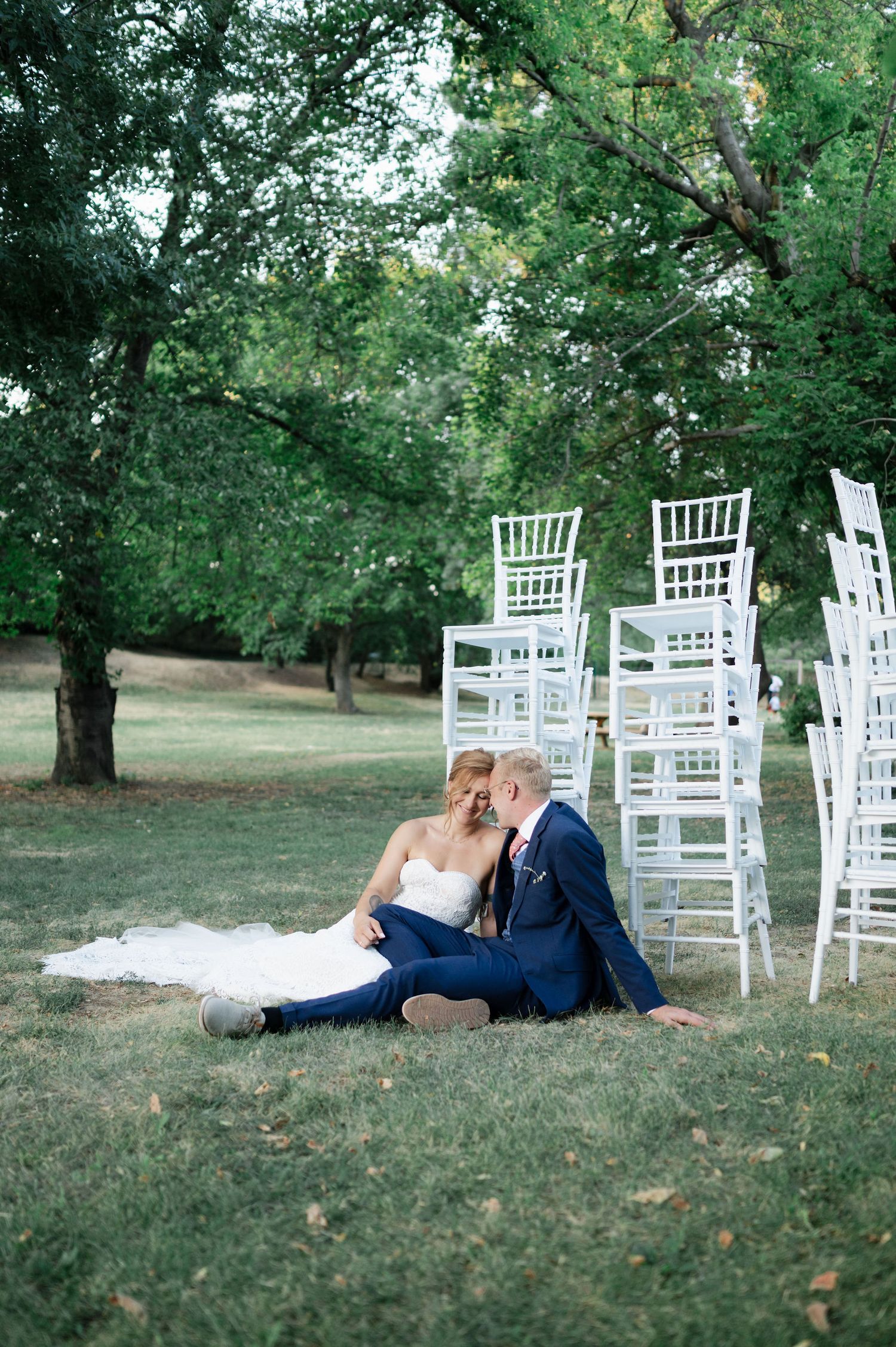 Couple in wedding attire laying on grass near white chairs in outdoor garden setting.