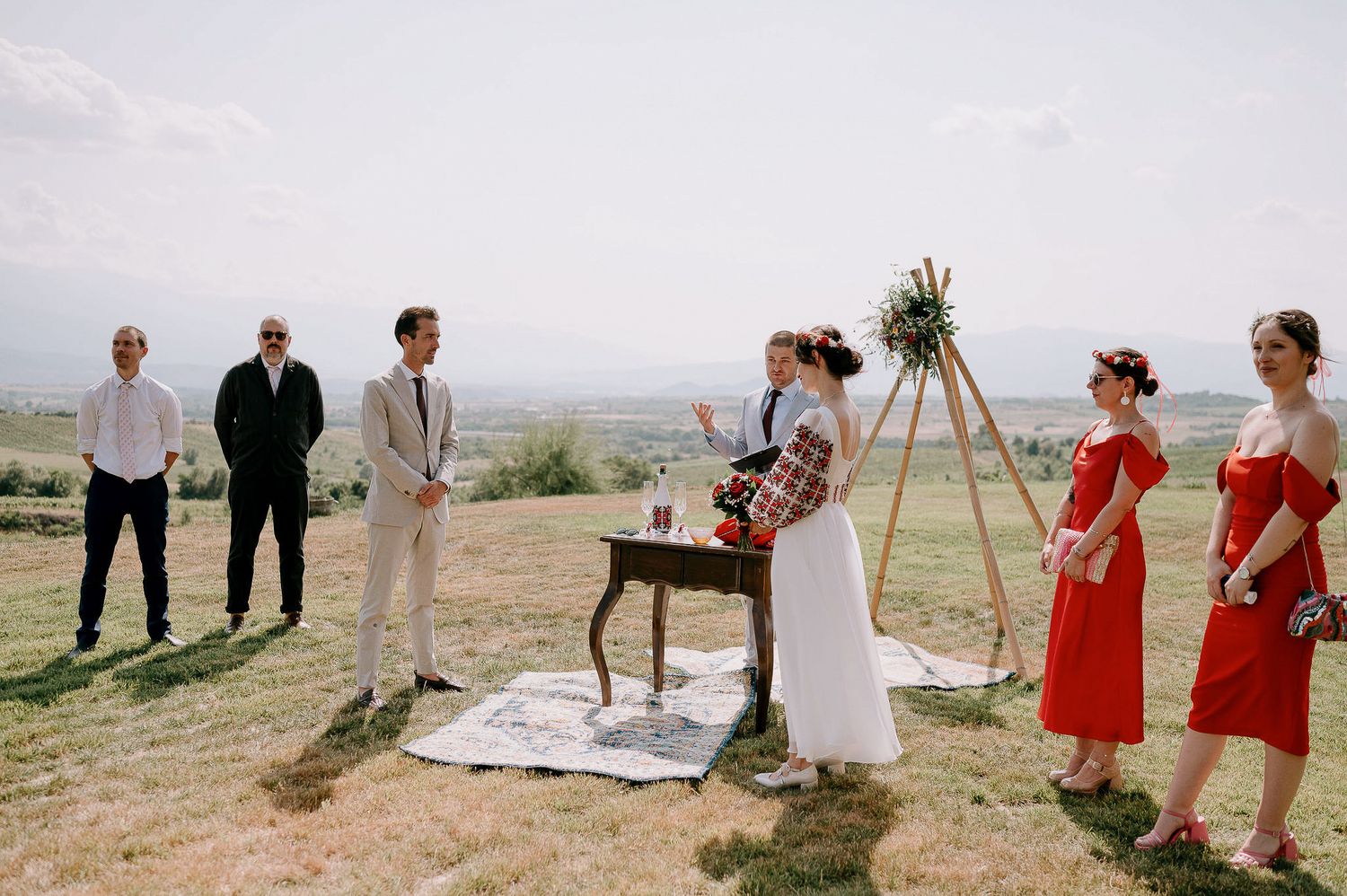 Outdoor wedding ceremony setup with wooden arch and red-dressed attendants on hillside.