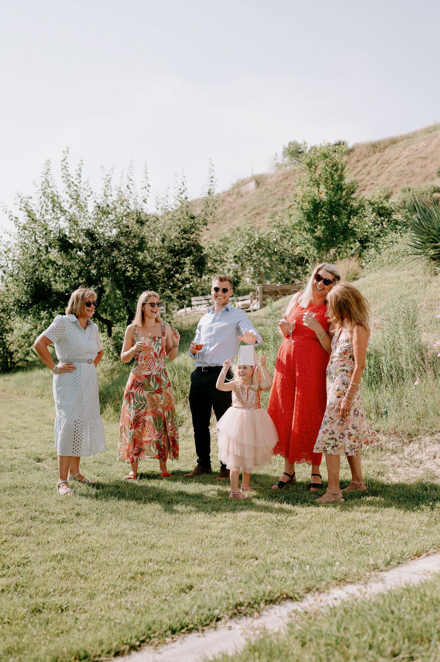 A group gathers on a grassy hillside wearing colorful summer dresses for a celebration.